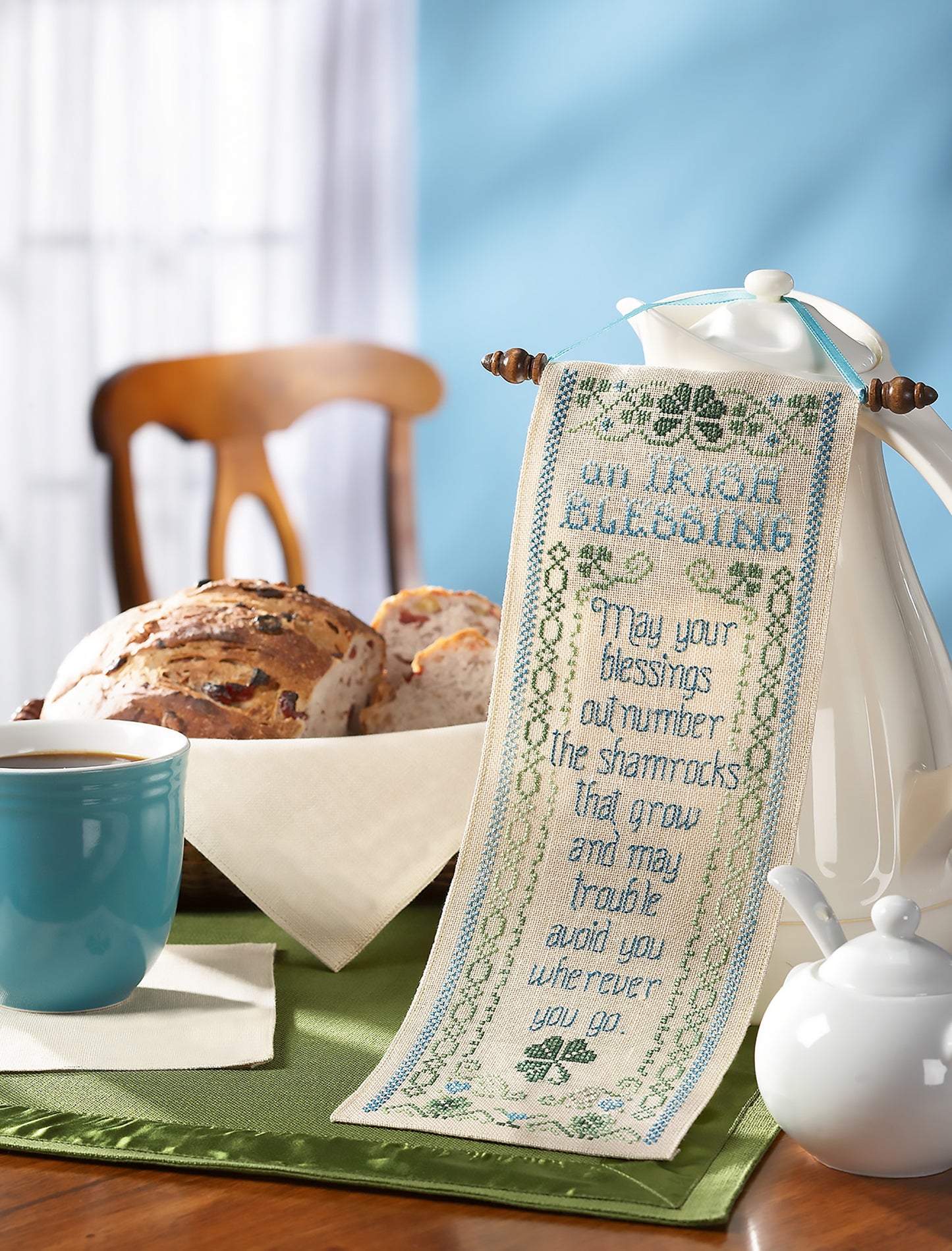 The Leisure Arts Special Sayings Cross Stitch hangs from a white teapot on a table with a blue mug, sliced bread, and green placemat, set against a wooden chair in a softly lit room—highlighting this charming friendship sampler.