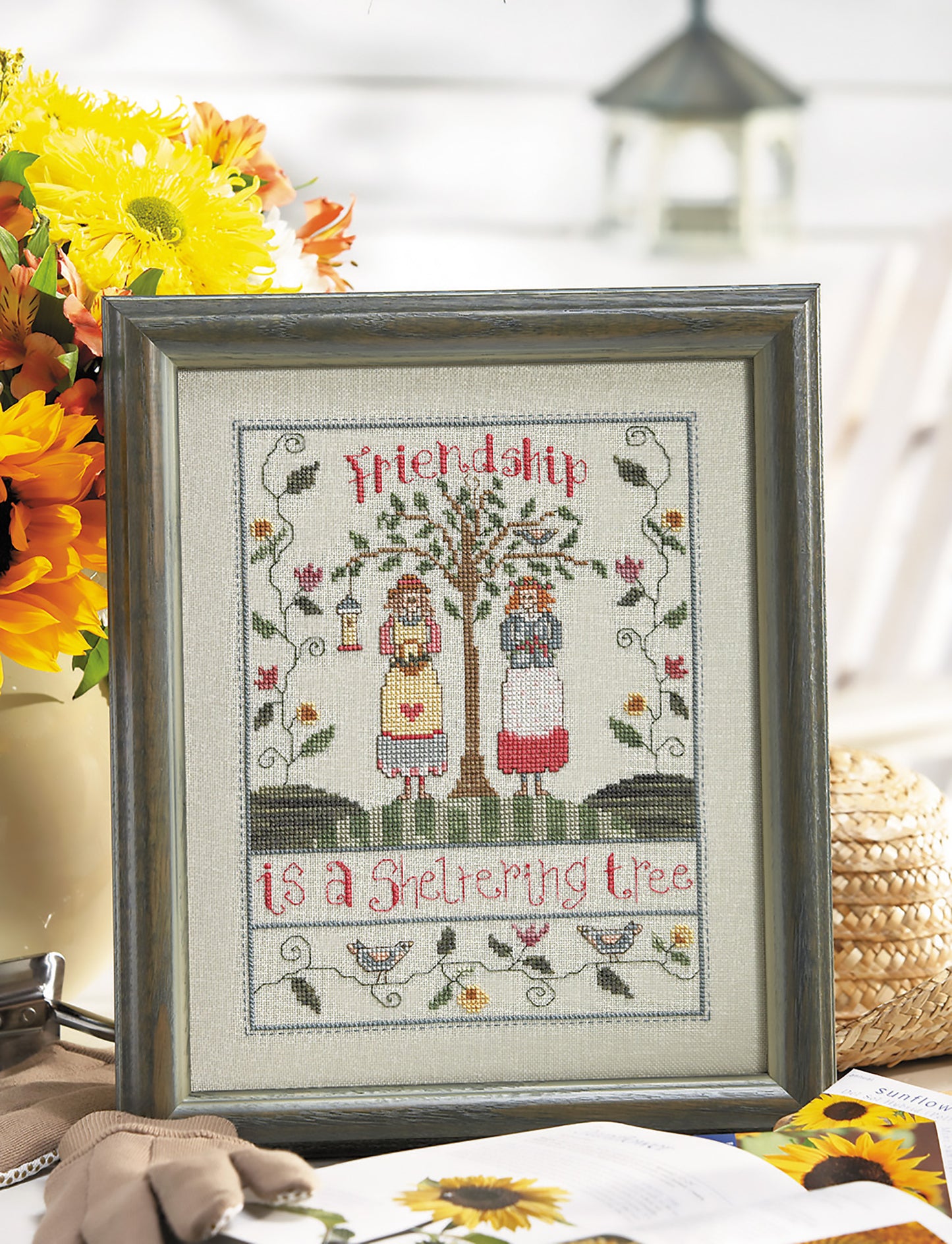 The Special Sayings Cross Stitch by Leisure Arts is a framed design showing two women beneath a tree with flowers and birds. “Friendship is a sheltering tree” appears on the artwork, alongside sunflowers and a straw hat.