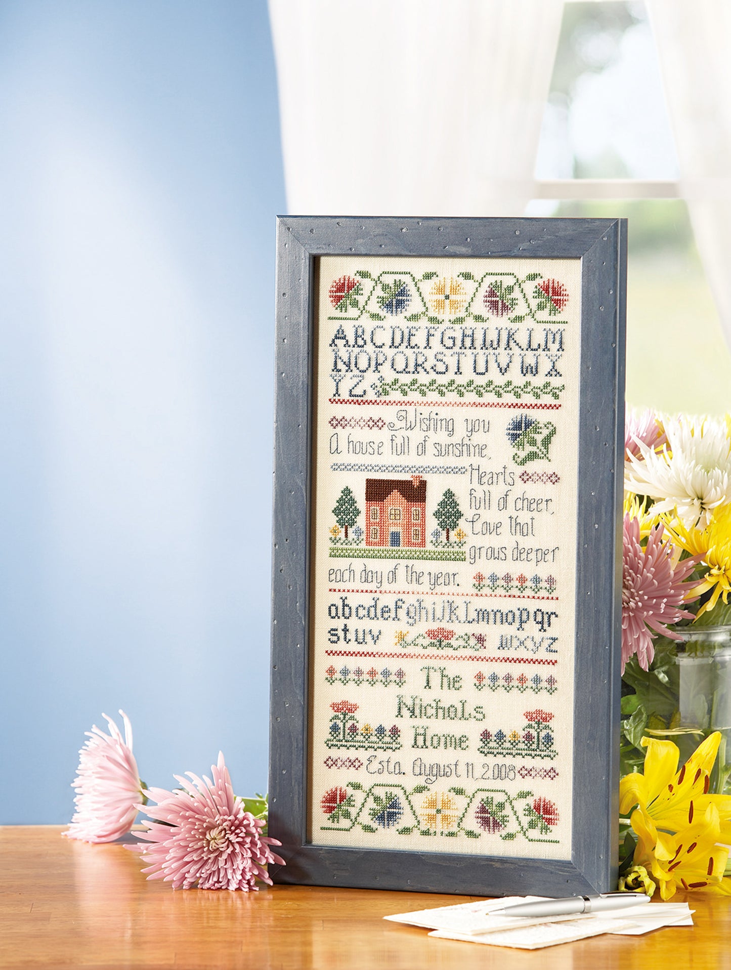 The Leisure Arts "Special Sayings Cross Stitch" sampler, framed and displayed with flowers, features the alphabet, a house, decorative patterns, and a welcoming message for the Nichols Home established on August 11, 2008.