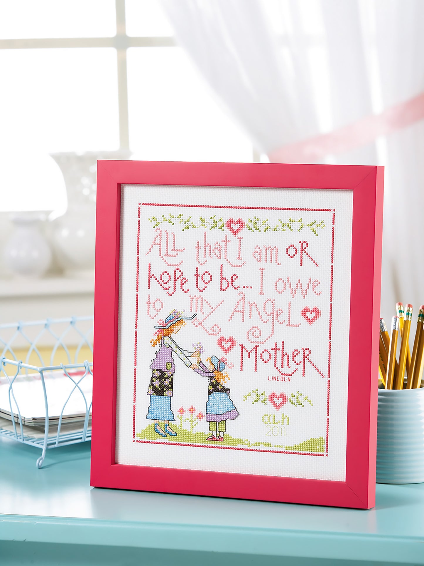 On a desk sits Leisure Arts’ Special Sayings Cross Stitch, a pink-framed piece reading, “All that I am or hope to be... I owe to my Angel Mother,” with a woman and child holding hands among flowers and hearts.
