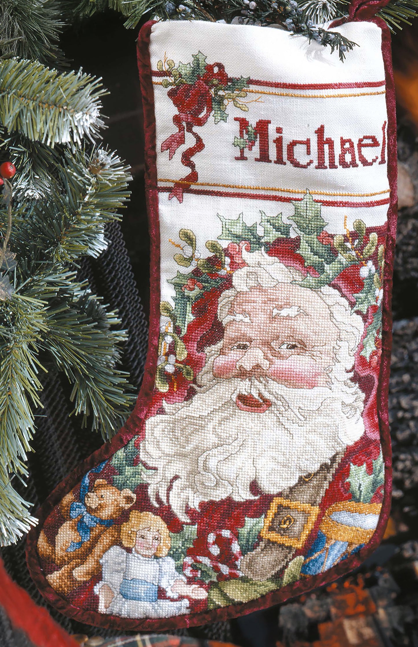 The Leisure Arts "Christmas Stocking—Create Heirlooms for Family and Friends" features a cross stitched “Michael,” Santa, holly leaves, and toys, displayed hanging on a Christmas tree among green pine branches.
