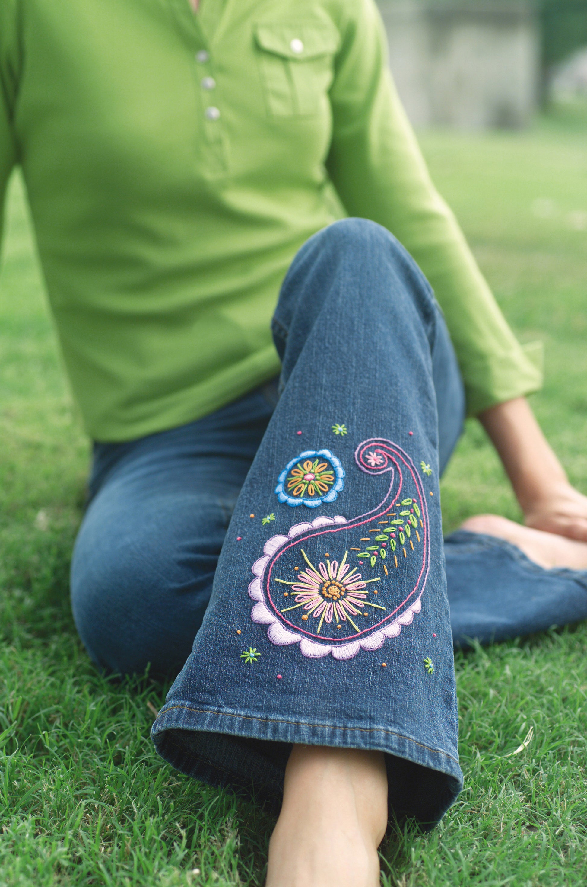 Seated cross-legged on grass, a person wears blue jeans with colorful embroidered floral and paisley hems—perfect inspiration for beginner projects from "Learn to Embroider with the Best of Leisure Arts" by Leisure Arts. Their face is not shown.