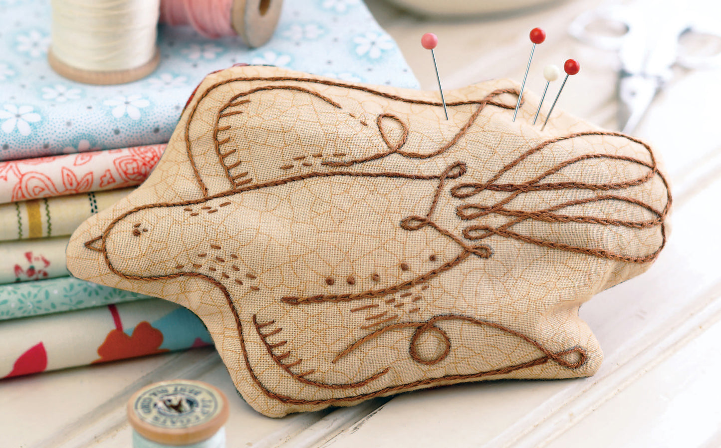 The Leisure Arts "Learn to Embroider with the Best of Leisure Arts" kit features a bird-shaped fabric pincushion with brown embroidery, perfect for beginners. It holds red-headed pins and sits on pastel fabrics beside sewing supplies.