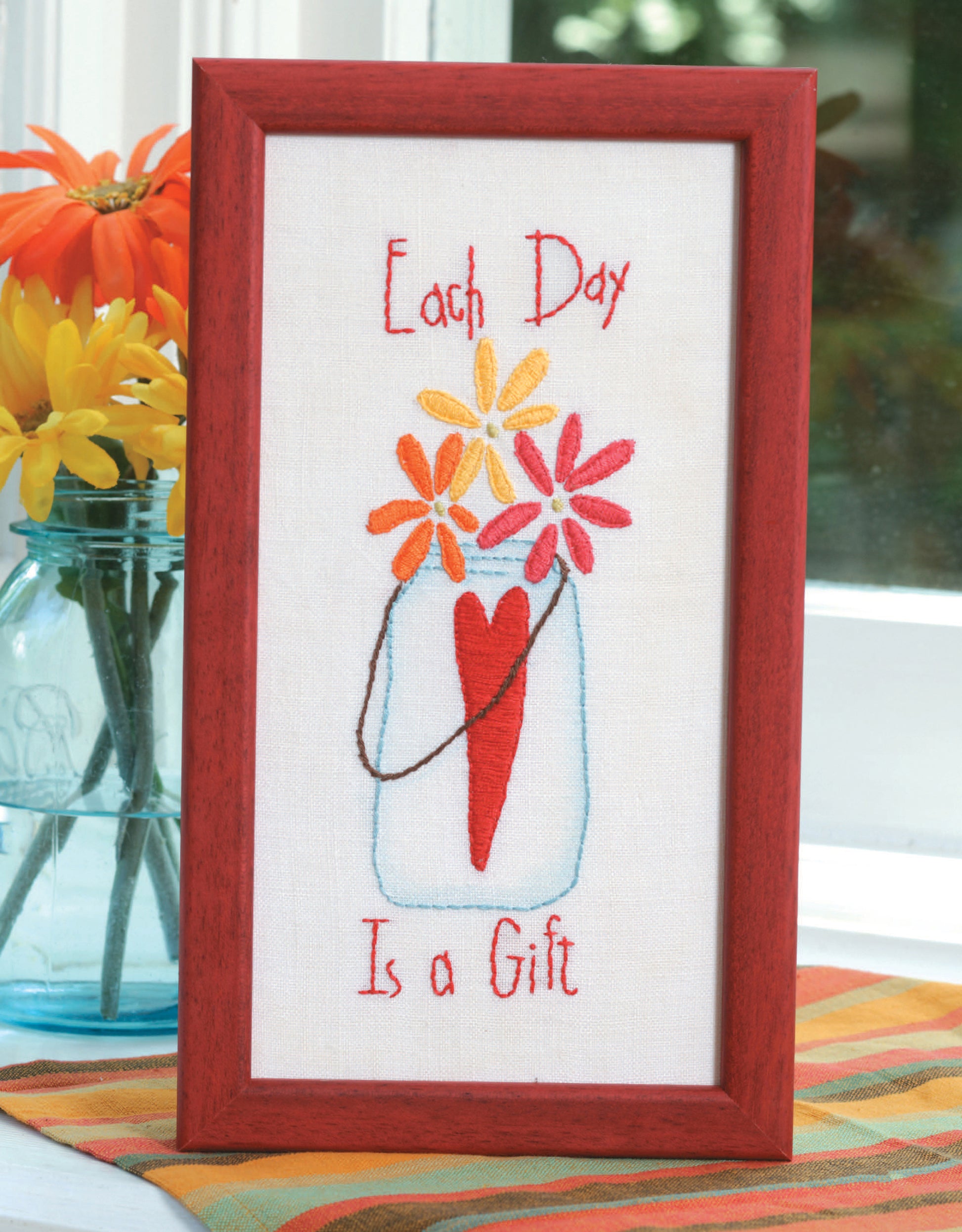 The Leisure Arts "Learn to Embroider with the Best of Leisure Arts" kit is perfect for beginners, featuring a mason jar of bright flowers and a red heart beside “Each Day Is a Gift” stitched in red.
