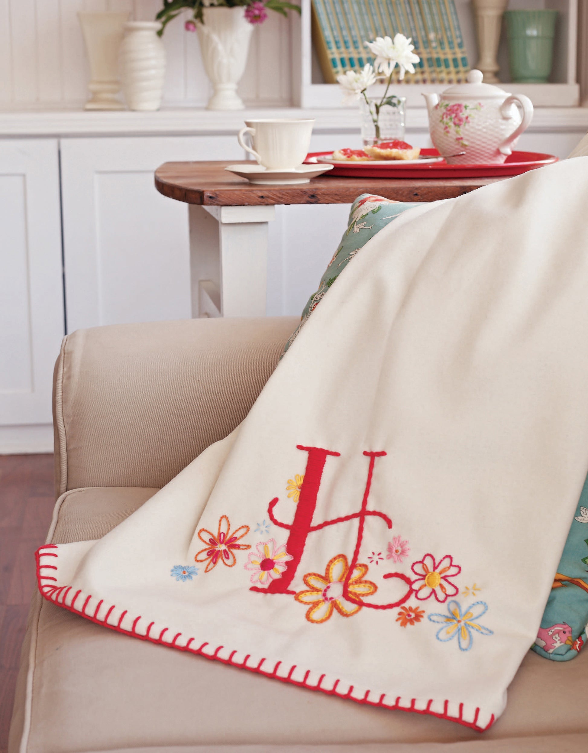 The book "Learn to Embroider with the Best of Leisure Arts" by Leisure Arts rests on a beige armchair, perfect for monogramming enthusiasts or beginner sewing projects. Nearby, a side table holds tea and snacks on a red tray.