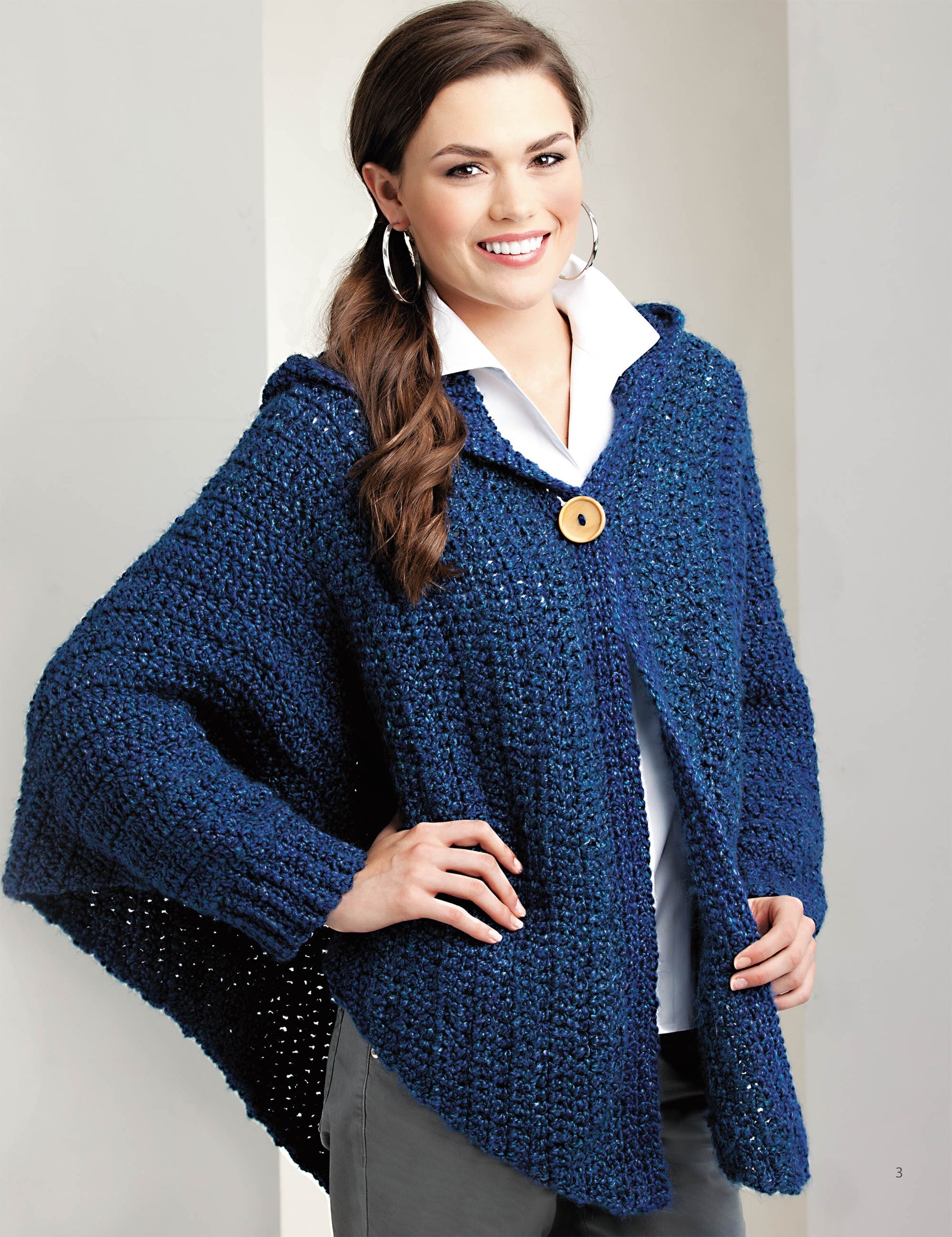 A woman smiles while wearing a chunky, dark blue knit poncho with a hood and large wooden button from Leisure Arts’ “Crochet the Cuffed Shawl & More”—perfect for those seeking easy patterns to craft cozy accessories.