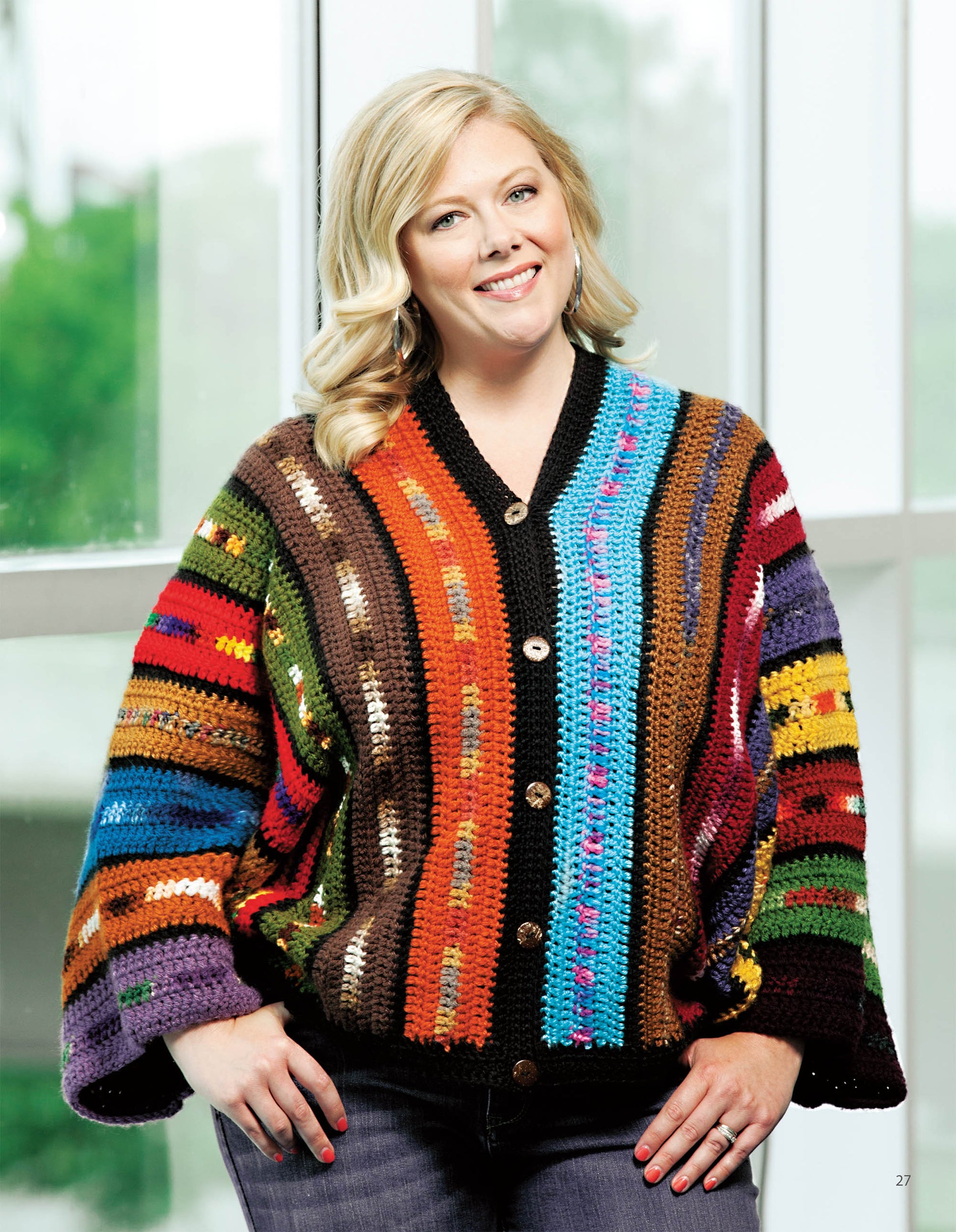 A smiling woman with blonde hair models the "Crochet the Cuffed Shawl & More" design by Leisure Arts, featuring a colorful, striped crochet cardigan with large buttons and a hybrid scarf, standing before sunlit windows with blurred greenery outside.