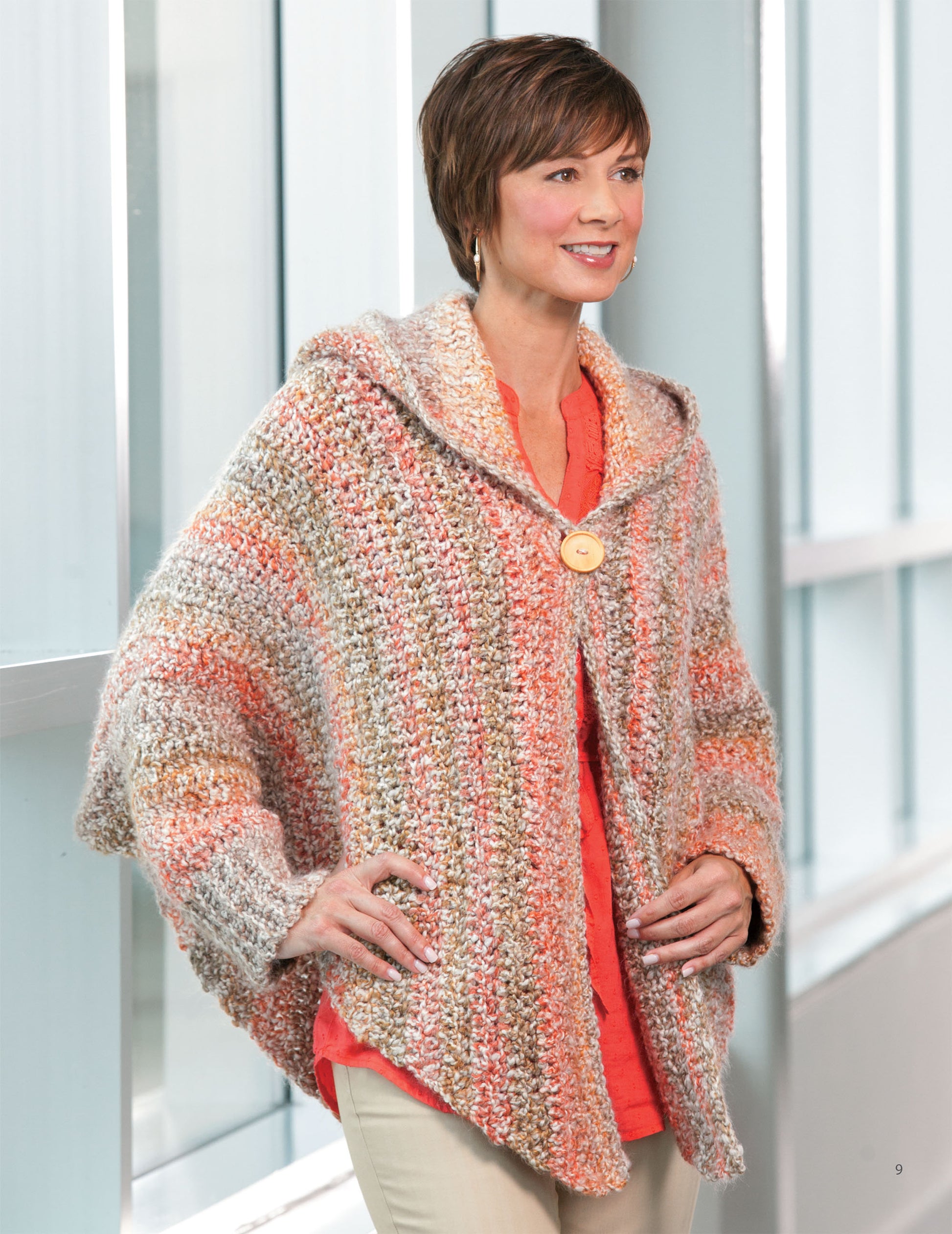 A woman with short brown hair, wearing a coral top and light pants, stands by a window smiling. She’s modeling the "Crochet the Cuffed Shawl & More" by Leisure Arts—a textured, multicolored knit poncho with a large collar button.