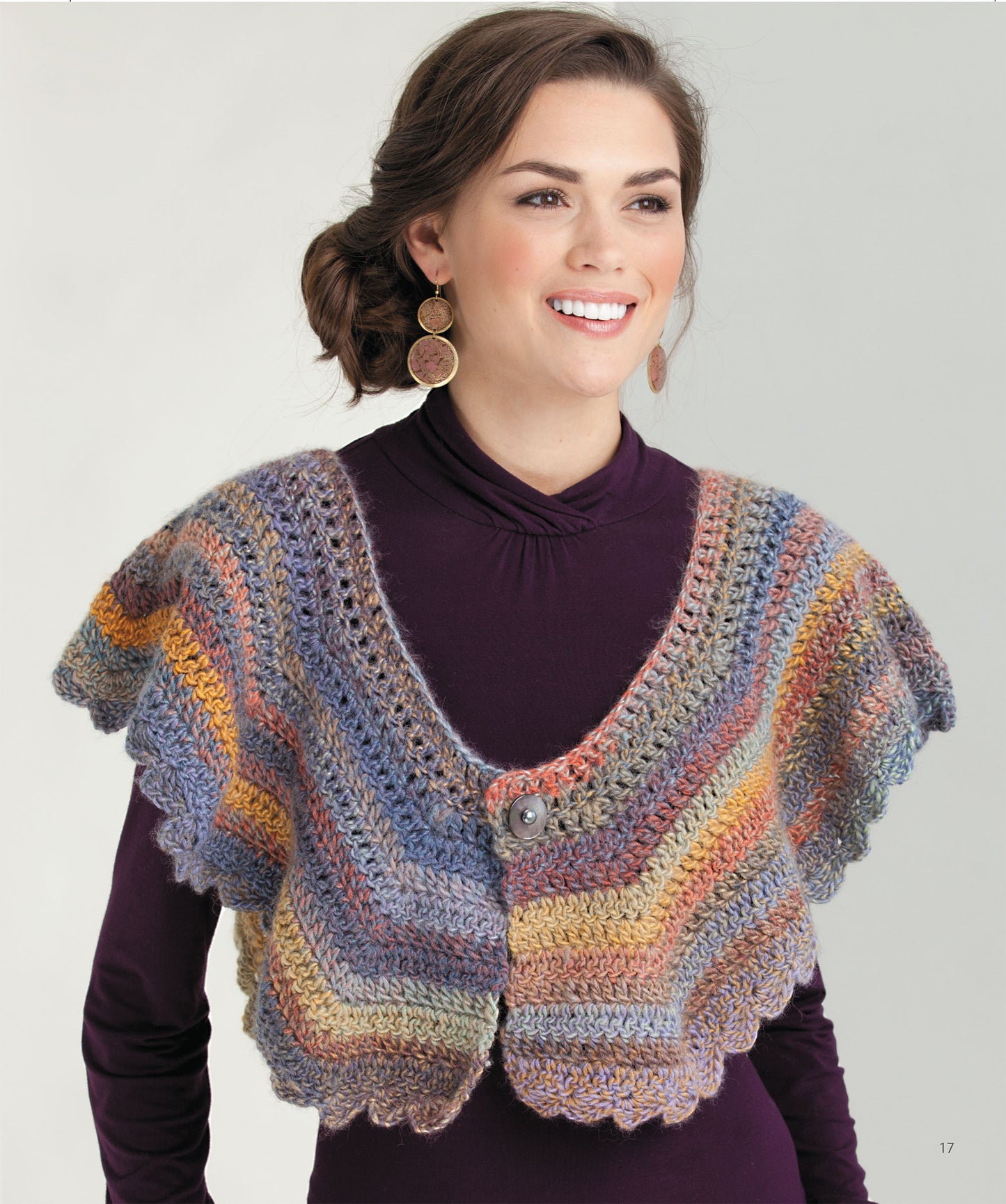 A woman with wavy brown hair wears circular earrings and a purple turtleneck while modeling the “Crochet the Cuffed Shawl & More” poncho-style capelet by Leisure Arts, featuring scalloped edges and a button closure.