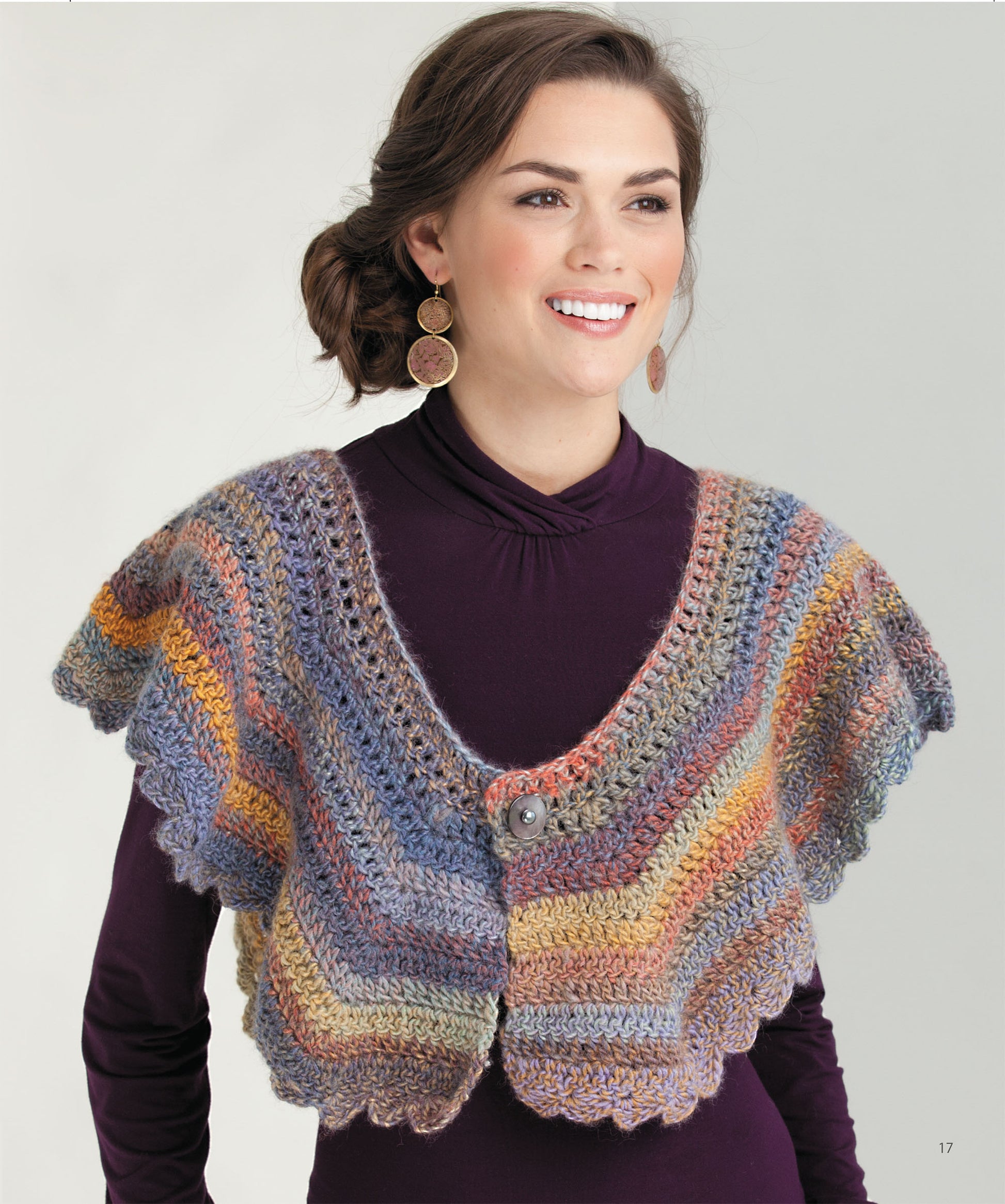 A woman with wavy brown hair wears circular earrings and a purple turtleneck while modeling the “Crochet the Cuffed Shawl & More” poncho-style capelet by Leisure Arts, featuring scalloped edges and a button closure.