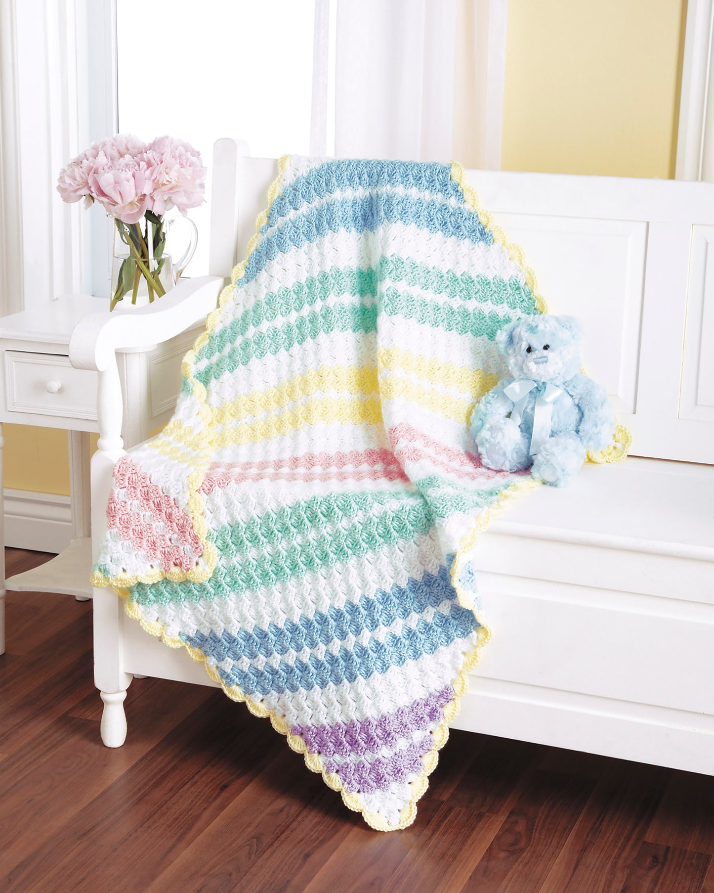 A bright, cozy room features a pastel-striped blanket from "Baby Afghans—The Best of Mary Maxim" by Leisure Arts, draped over a white bench with a blue teddy bear nearby and pink flowers in a vase on the side table.