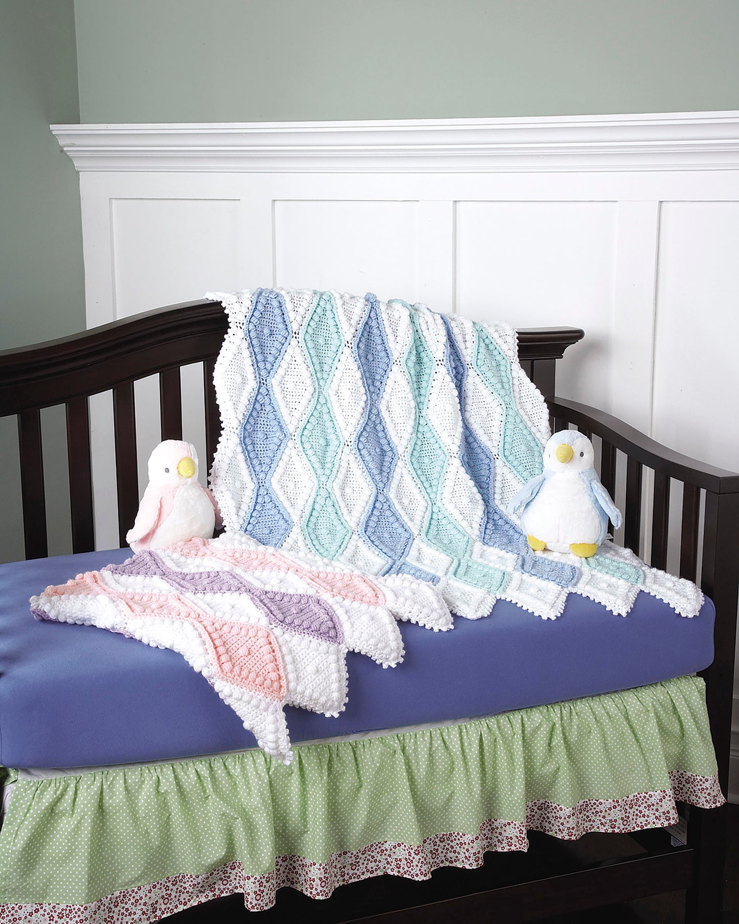 A crib with a purple mattress and green bed skirt displays two pastel baby afghans from "Baby Afghans— The Best of Mary Maxim" by Leisure Arts, draped over the side, plus two plush penguin toys—one pink and one blue.