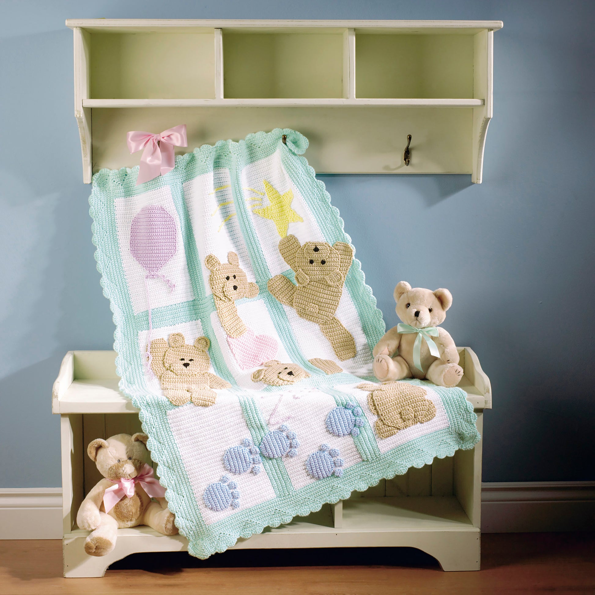 On a white bench, two teddy bears sit next to a pastel crocheted afghan from "Baby Afghans—The Best of Mary Maxim" by Leisure Arts. Above, a pink ribbon hangs against a light blue wall—ideal inspiration for beginner crochet projects.