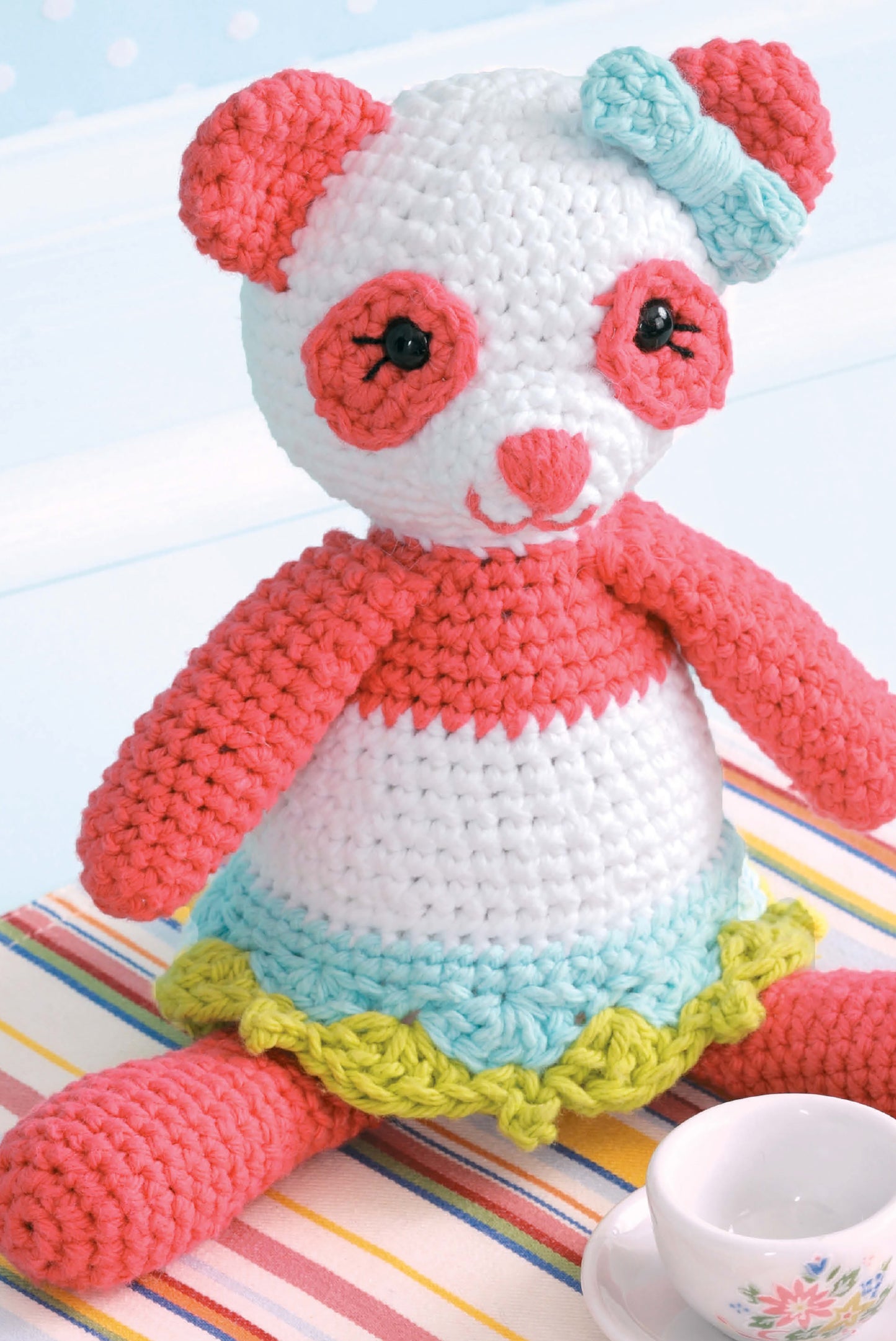 A crocheted panda toy from Leisure Arts' "Learn to Crochet Toys" features a pink and white body, blue bow, and frilled skirt with blue and yellow ruffles—an adorable project for beginner crocheters.