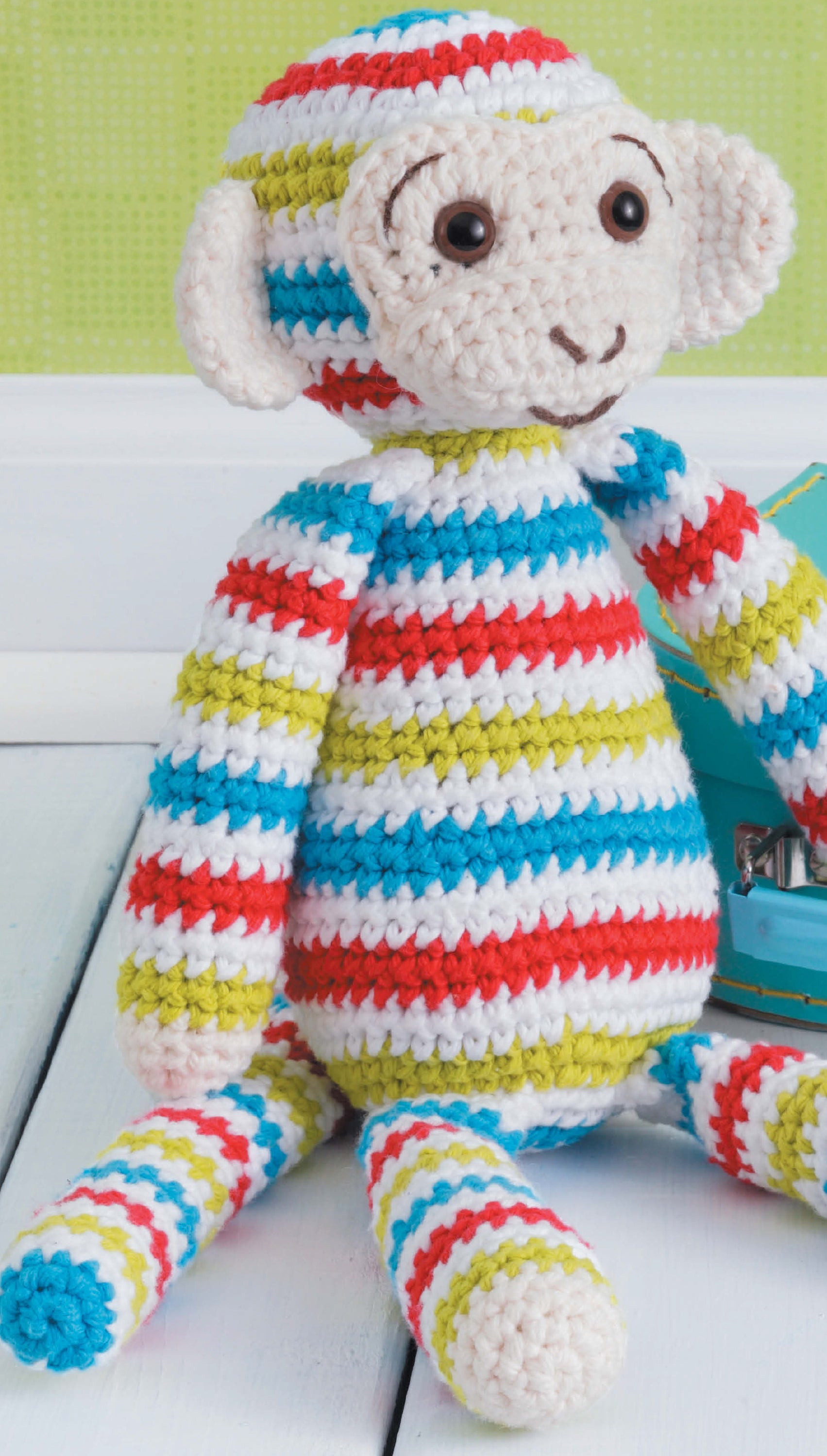 The Leisure Arts Learn to Crochet Toys kit lets you make a striped amigurumi monkey in red, blue, green, yellow, and white—smiling with embroidered features. Perfect for beginners, it’s pictured on a light wood floor against a soft green background.