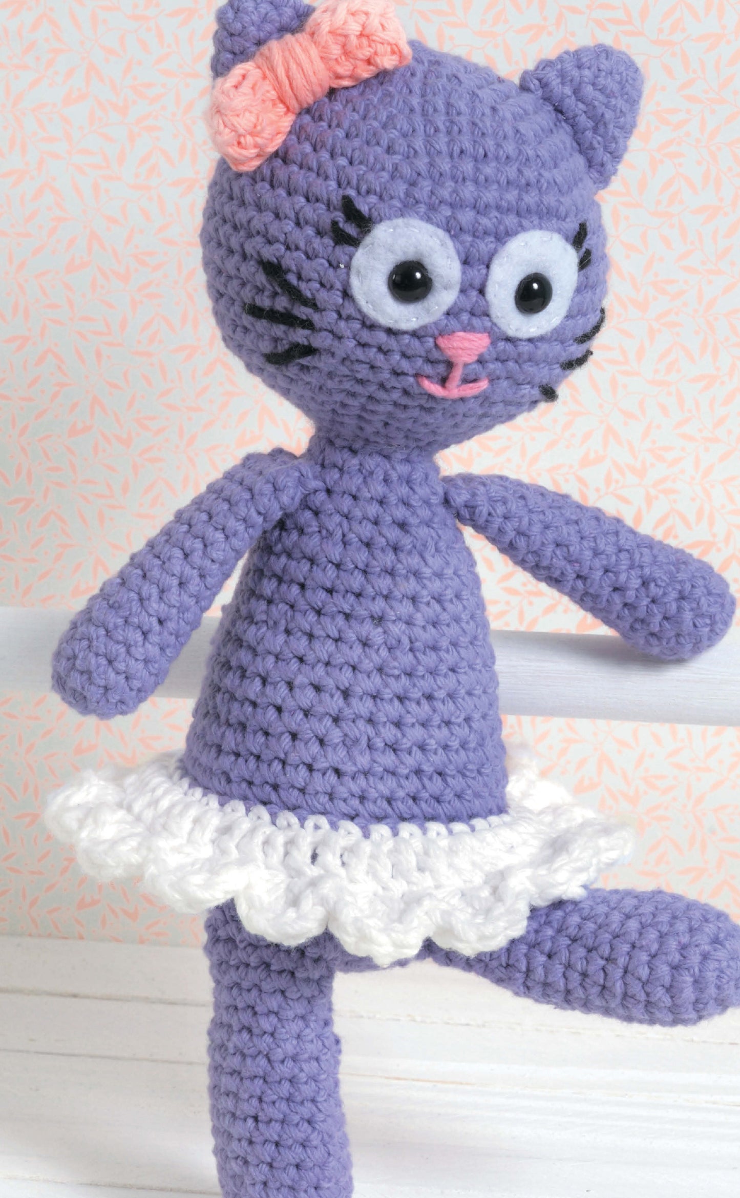 The Leisure Arts "Learn to Crochet Toys" kit features a charming amigurumi purple cat doll with large googly eyes, pink bow, black whiskers, pink nose and mouth, and a white ruffled skirt—ideal for beginner crochet projects.