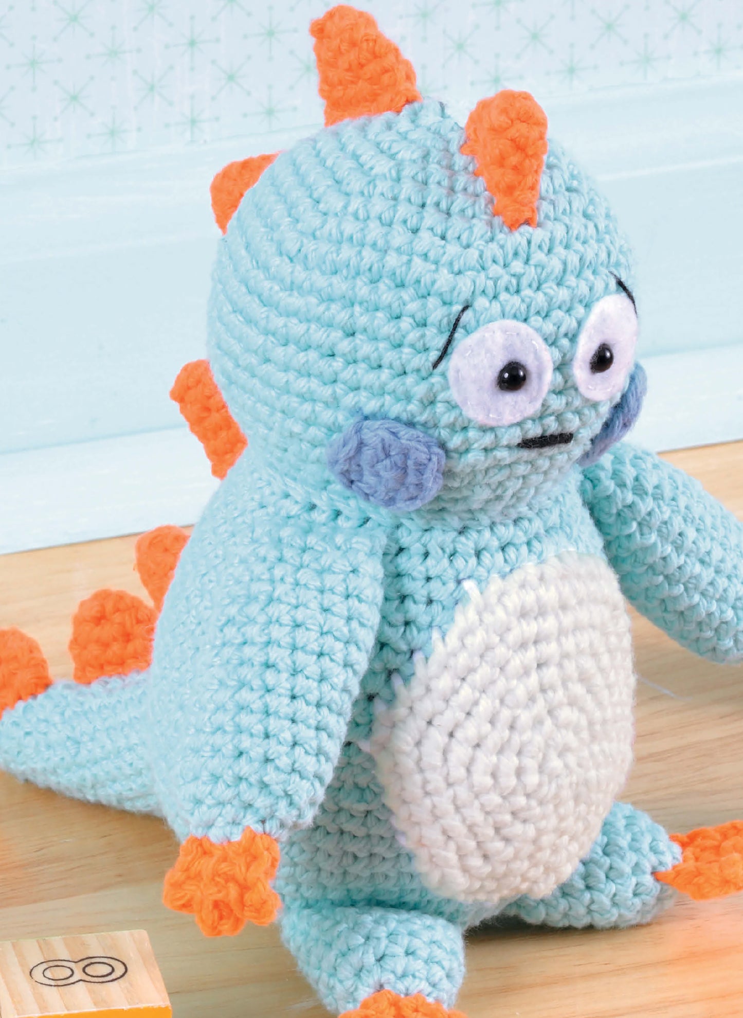The "Learn to Crochet Toys" kit by Leisure Arts features a light blue amigurumi dinosaur with orange spikes, big round eyes, blue cheeks, and orange feet and hands, shown sitting on a wooden floor near a toy block.