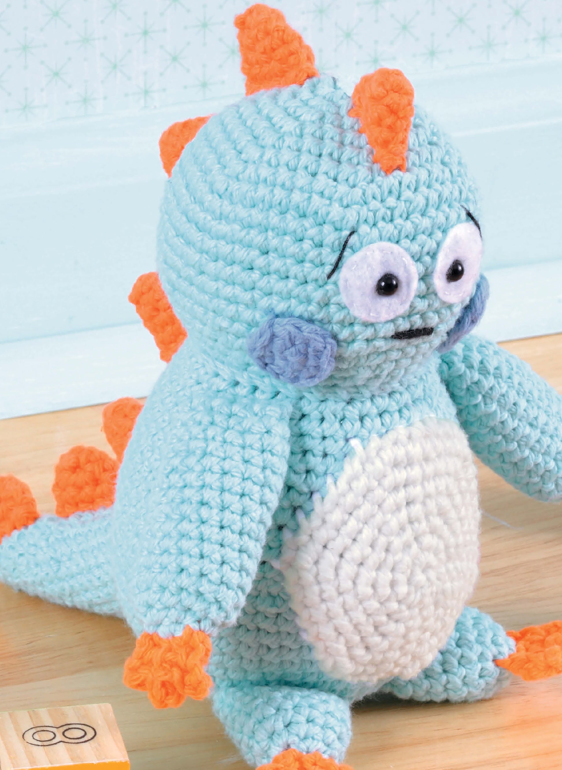 The "Learn to Crochet Toys" kit by Leisure Arts features a light blue amigurumi dinosaur with orange spikes, big round eyes, blue cheeks, and orange feet and hands, shown sitting on a wooden floor near a toy block.