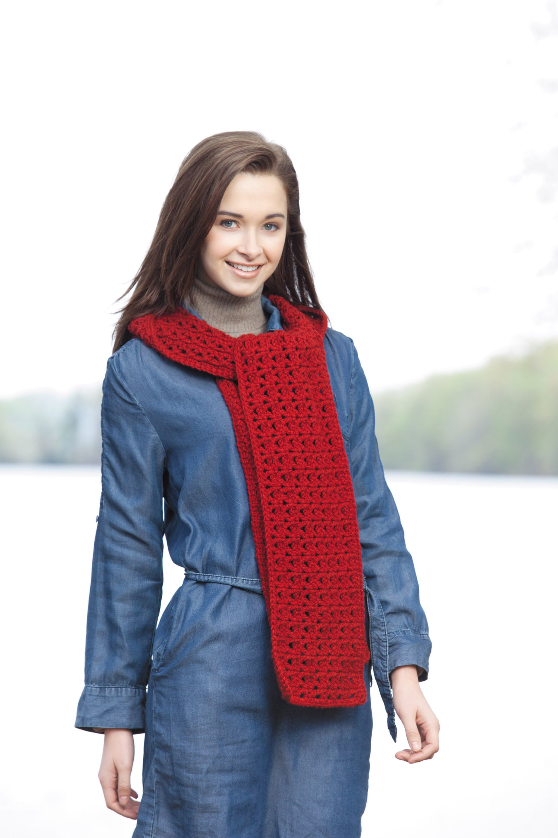 A young woman with brown hair wears a denim jumpsuit and a bright red crocheted scarf from Leisure Arts’ Hooded Scarves, Book 2 Digital Download, standing outdoors with a blurred background of trees and water.