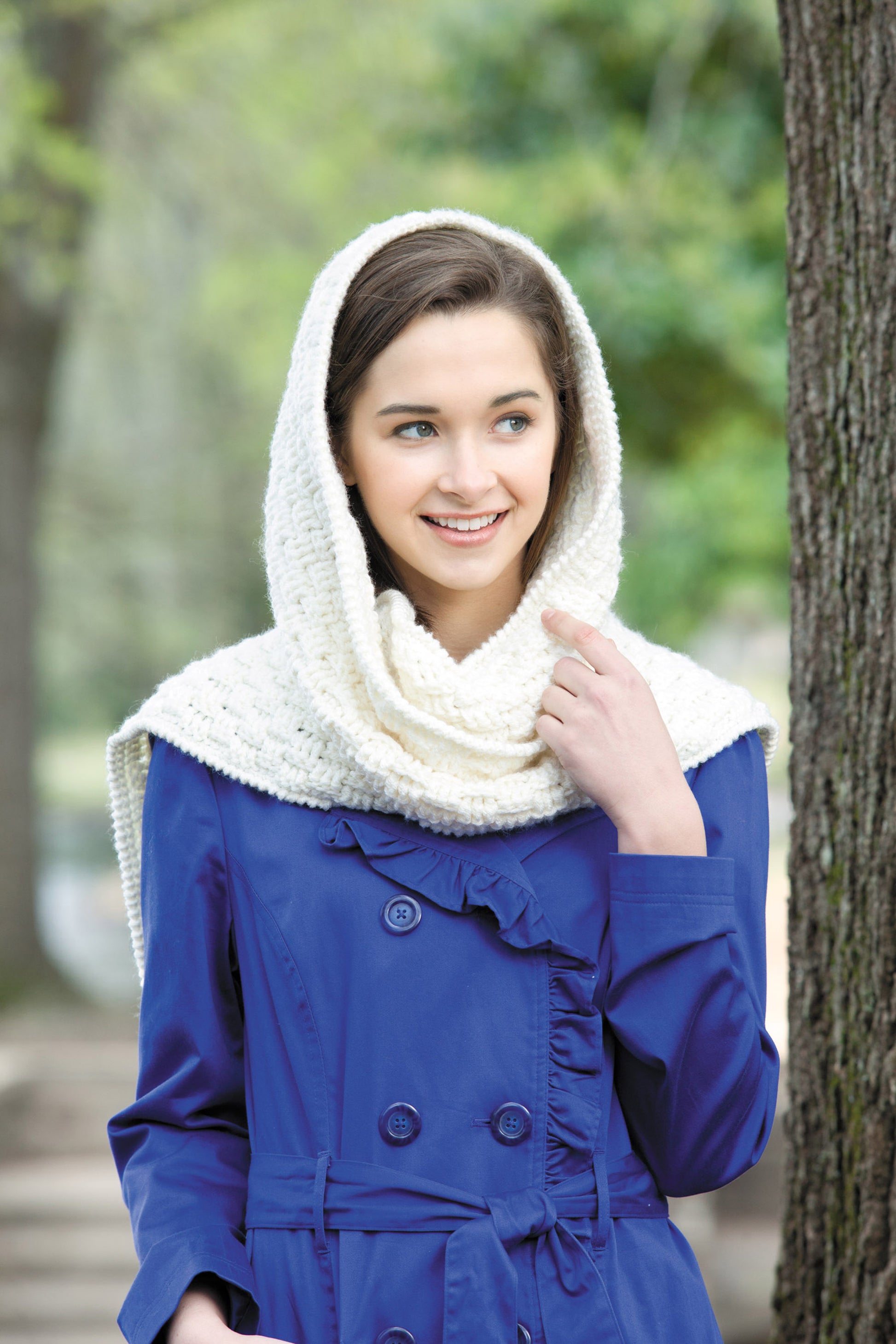 A woman wears a blue coat and a hooded cream-colored scarf, smiling outdoors. Her look features cozy inspiration from "Hooded Scarves, Book 2 Digital Download" by Leisure Arts—perfect for easy or beginner crochet projects.