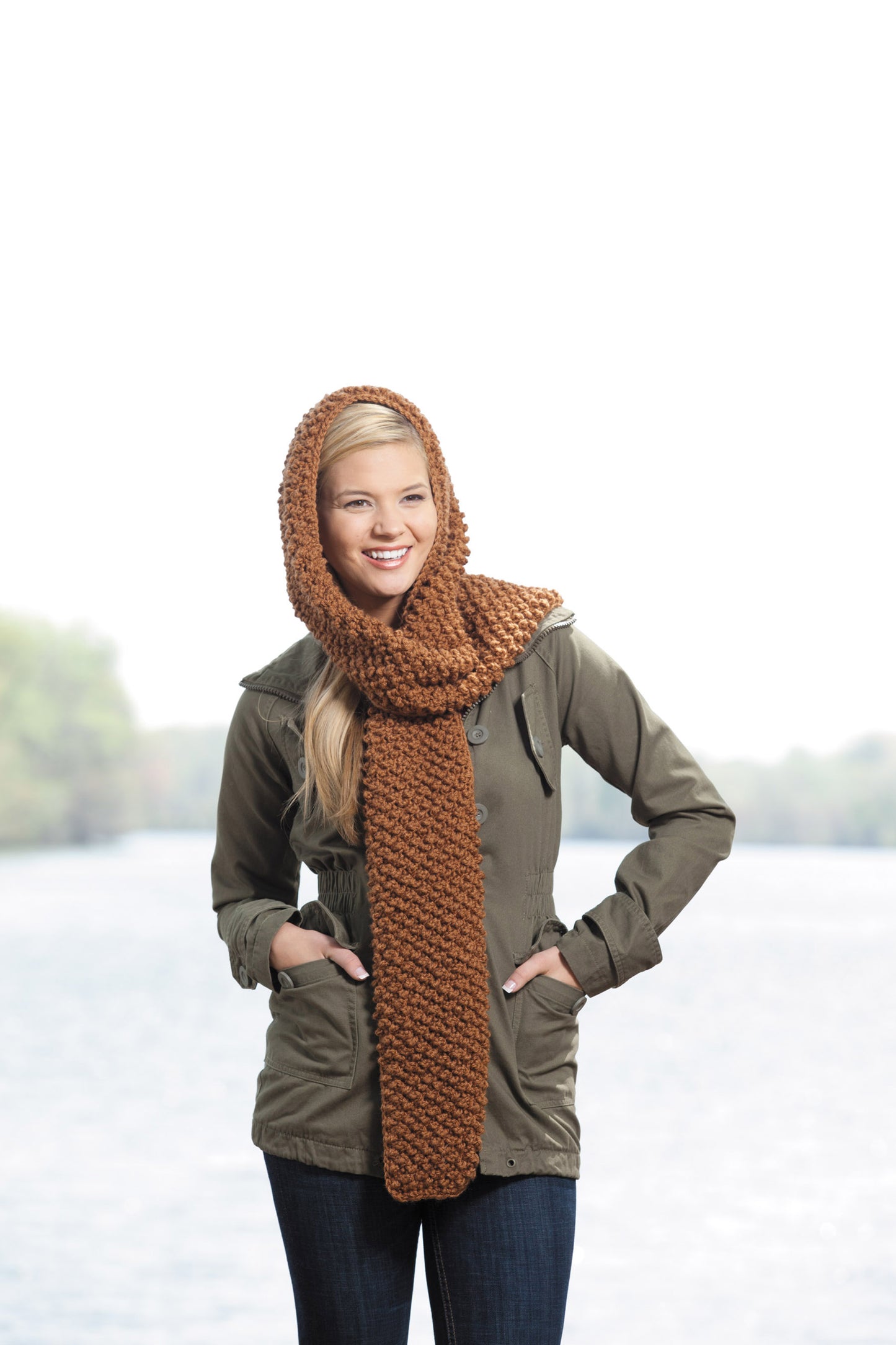 Wearing a cozy green jacket and a chunky brown crochet scarf, a woman smiles by the water. Get inspired with "Hooded Scarves, Book 2 Digital Download" from Leisure Arts for your next stylish crochet project!.