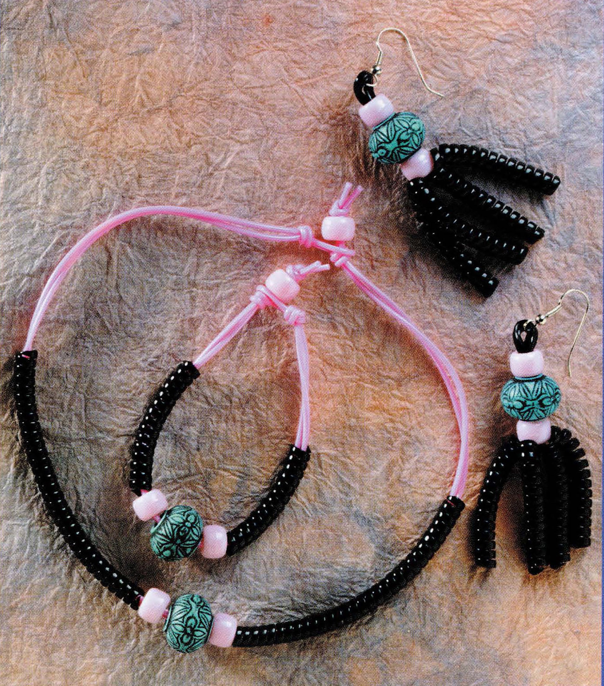 The Leisure Arts Plastic Lacing & Pony Beads set includes a handmade necklace, bracelet, and earrings in pink cord with black and floral beads, stylishly arranged on a textured surface.