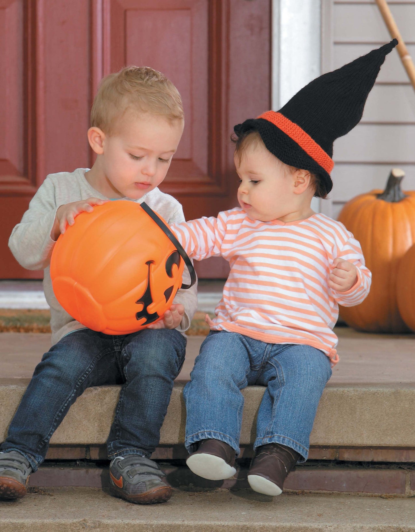 Two young children sit on porch steps. One holds an orange pumpkin bucket, while the other, wearing a Leisure Arts Knit Hat for Babies and a striped shirt, reaches for it. A pumpkin is nearby by the red door.