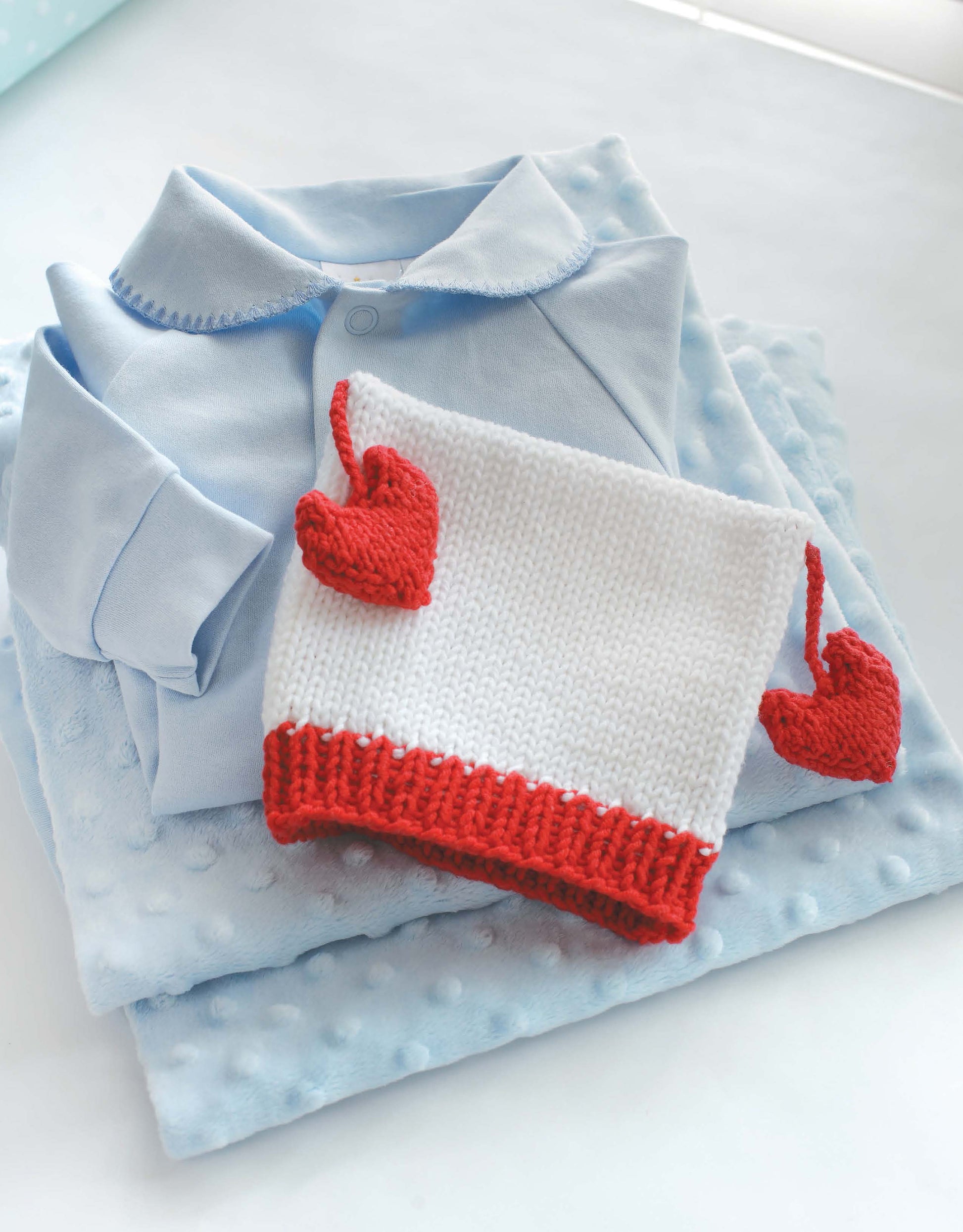 A folded light blue baby onesie lies on a soft blue blanket. Topping it is a Leisure Arts Knit Hats for Babies—a white knit hat with red edges and two red hearts—adding extra charm to the scene.