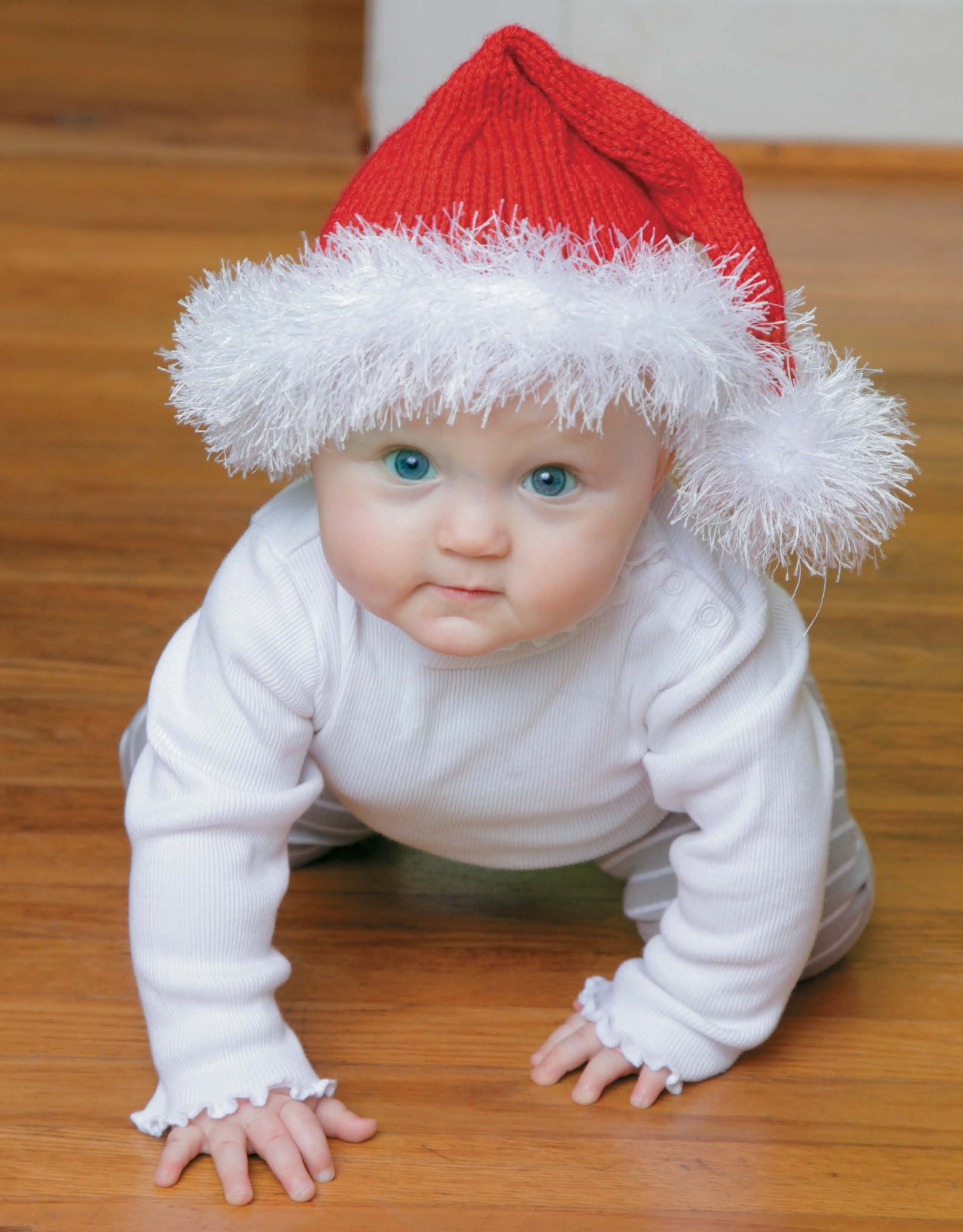 A baby with blue eyes crawls on a wooden floor, looking up at the camera while wearing a red and white Knit Hat for Babies by Leisure Arts and a white outfit.