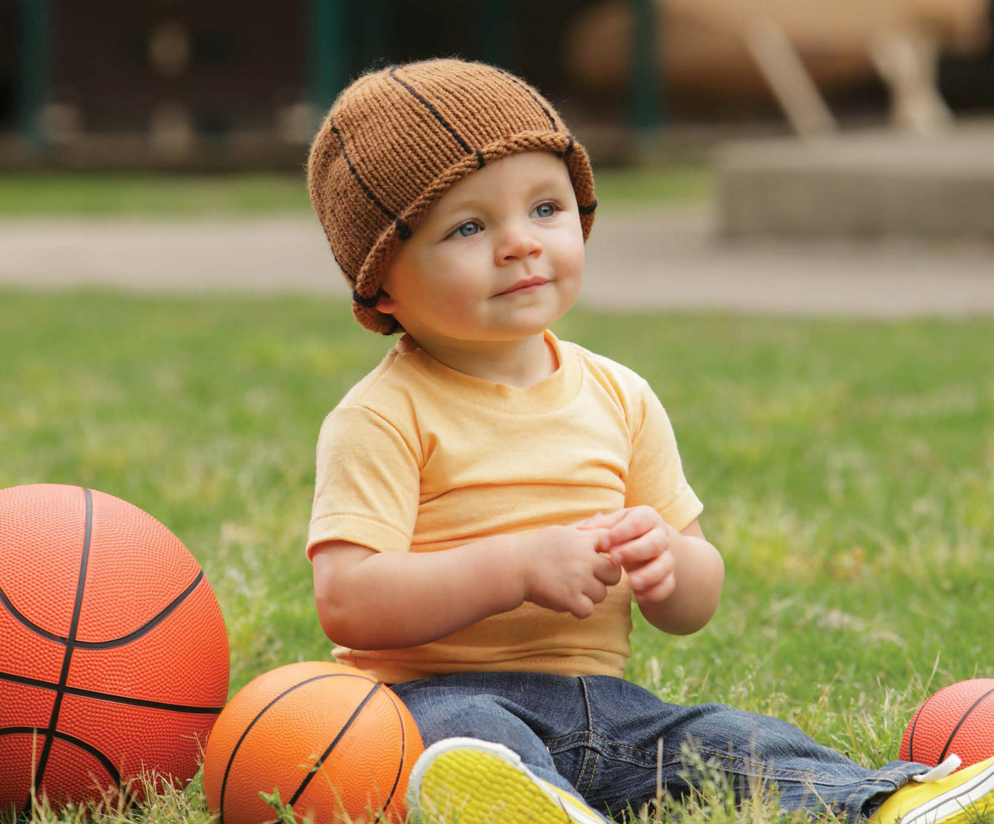 A young child wears a Leisure Arts Knit Hat for Babies, paired with a yellow t-shirt and jeans, as they sit on grass surrounded by three differently sized basketballs, smiling and glancing to the side.