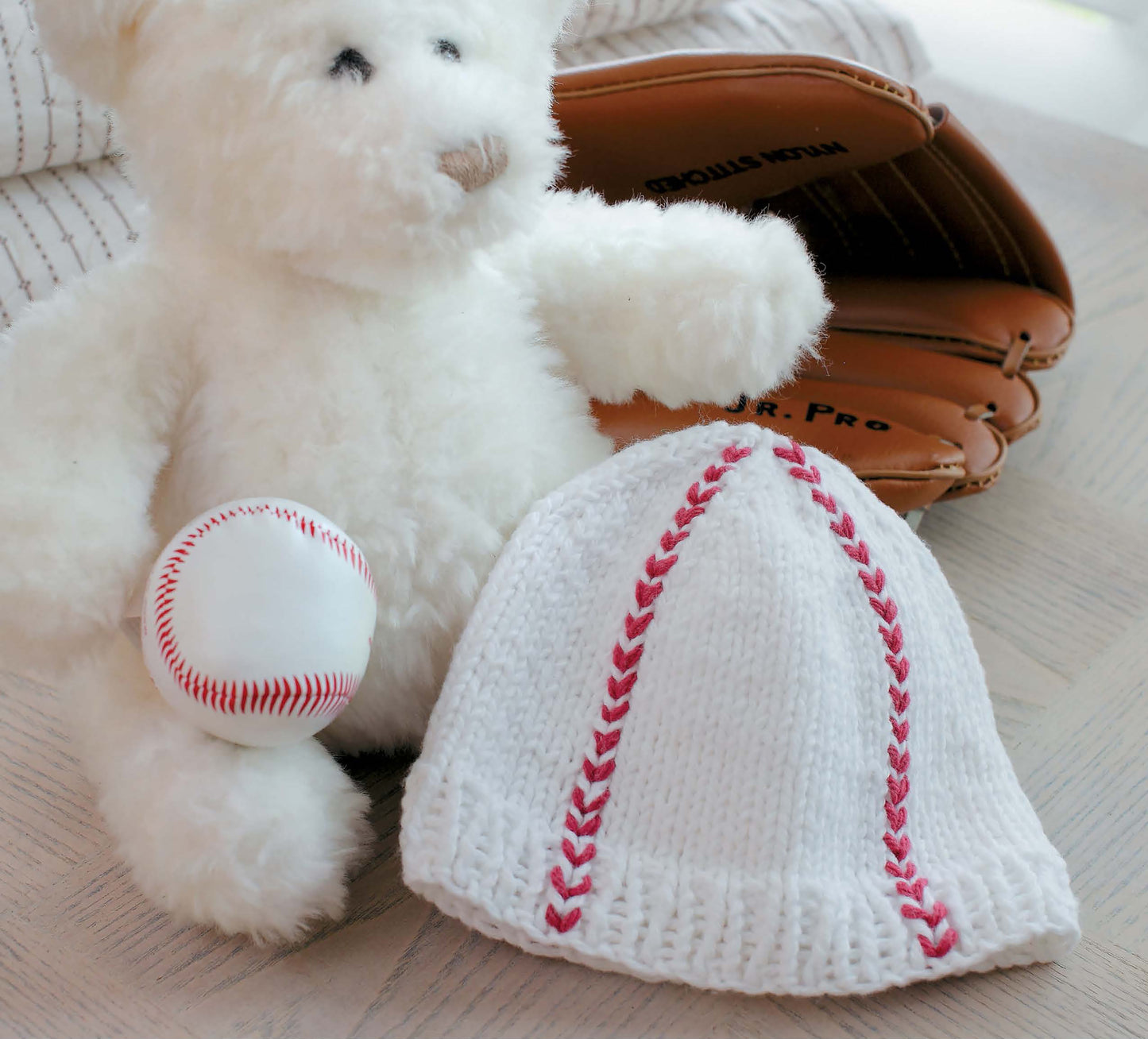 A white teddy bear holds a small baseball while wearing a cozy baby knit hat with red stitching, from "Knit Hats for Babies" by Leisure Arts. A brown glove sits behind them, all on a light wooden surface—great knitting inspiration!.