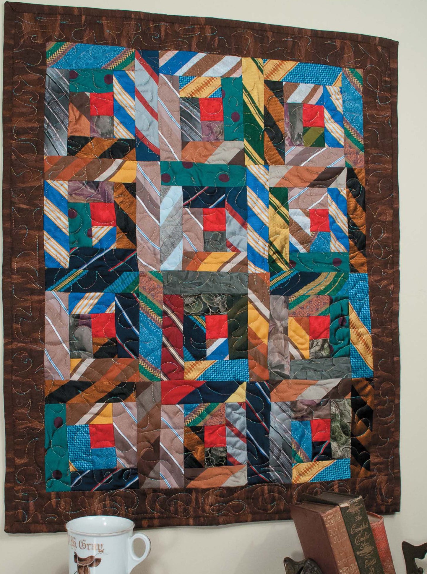 The "Memory Quilts—Create Treasured Heirlooms for Loved Ones!" by Leisure Arts features geometric patchwork in blue, red, orange, and green with a brown border—perfect above a table with books and a mug, inspired by beginner quilt patterns.