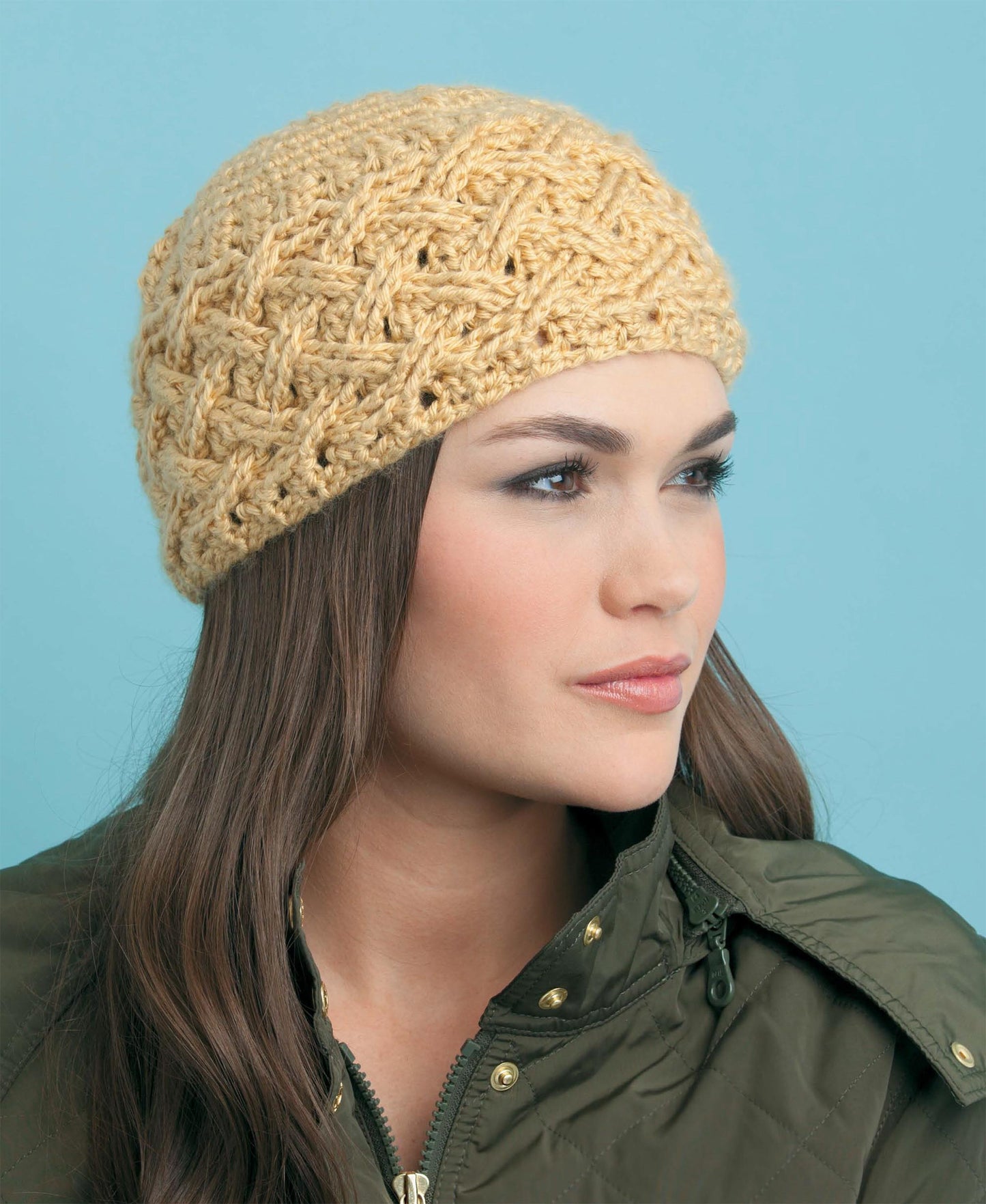 A woman with long brown hair wears a textured, light yellow crochet hat inspired by cable techniques from "Beginner's Guide to Crochet Cables" by Leisure Arts, paired with an olive quilted jacket against a light blue background.