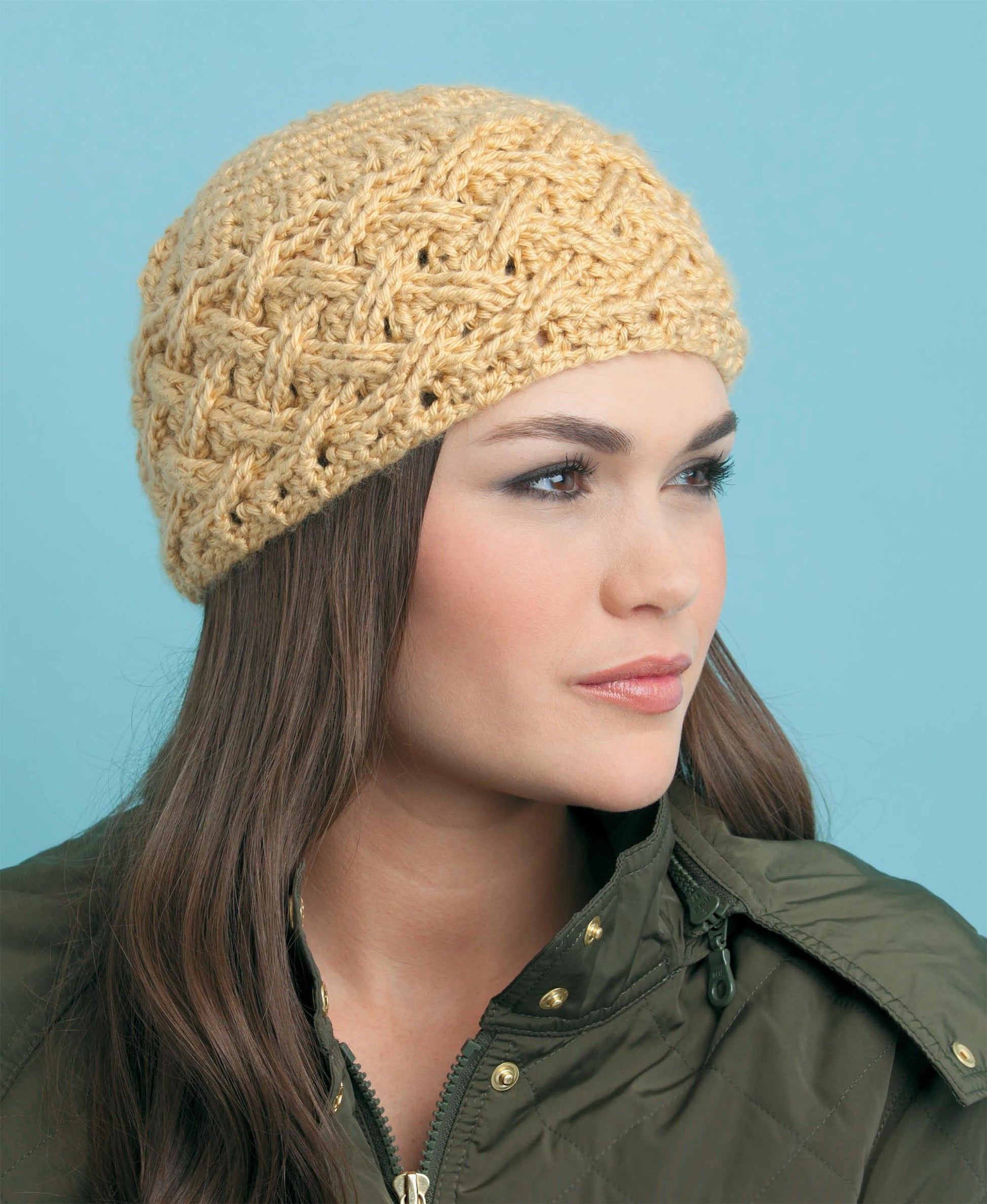 A woman with long brown hair wears a textured, light yellow crochet hat inspired by cable techniques from "Beginner's Guide to Crochet Cables" by Leisure Arts, paired with an olive quilted jacket against a light blue background.