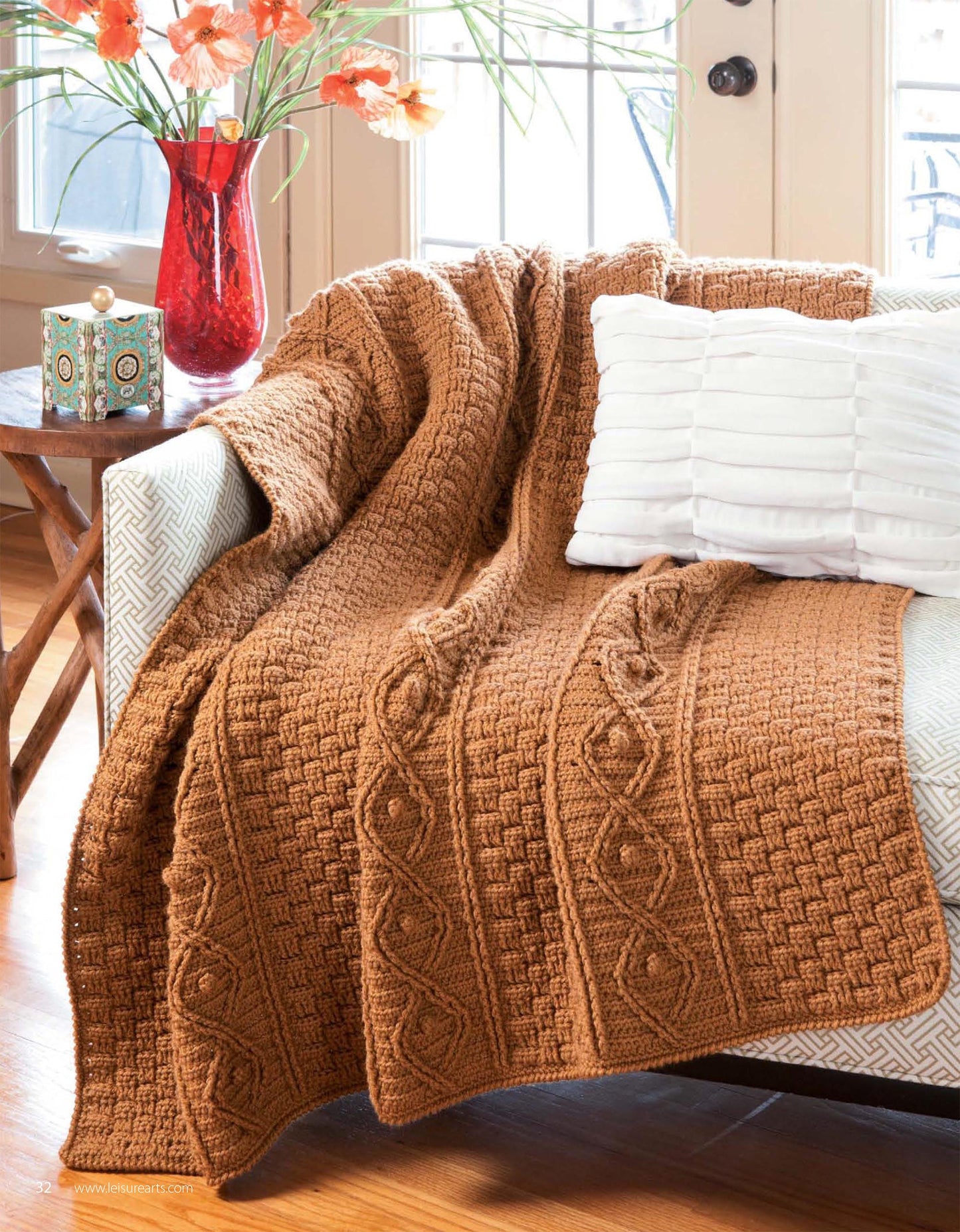 The book "Beginner's Guide to Crochet Cables" by Leisure Arts rests on a wooden table beside a beige armchair with a white pillow, red vase of orange flowers, and decorative box in a sunlit room.