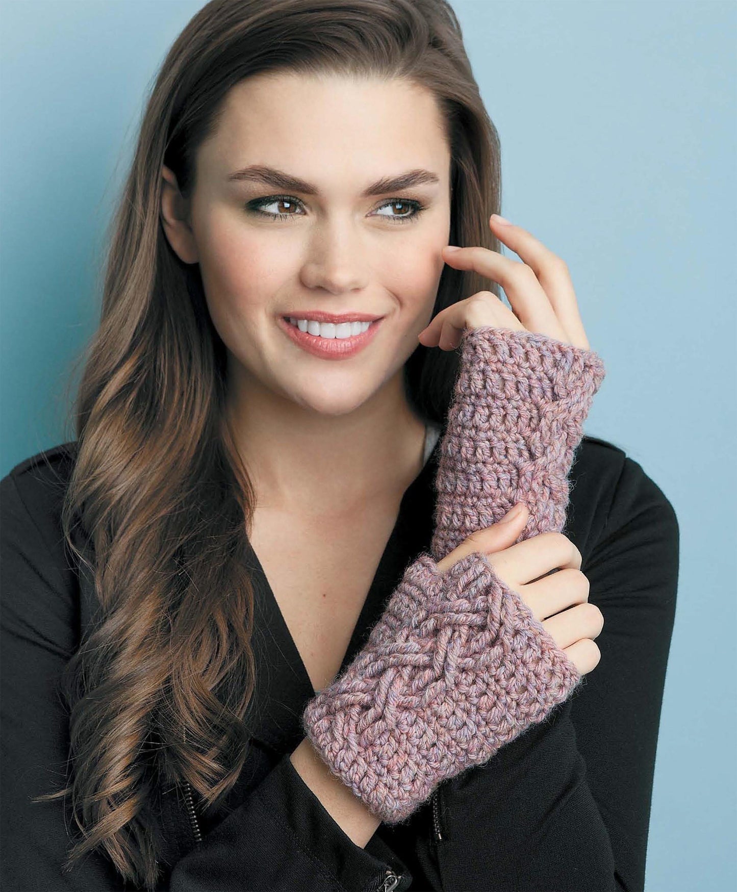 A smiling woman with long brown hair wears a black top and displays chunky, hand-knitted light purple fingerless gloves with crochet cable patterns, made using Leisure Arts' Beginner's Guide to Crochet Cables, against a light blue background.