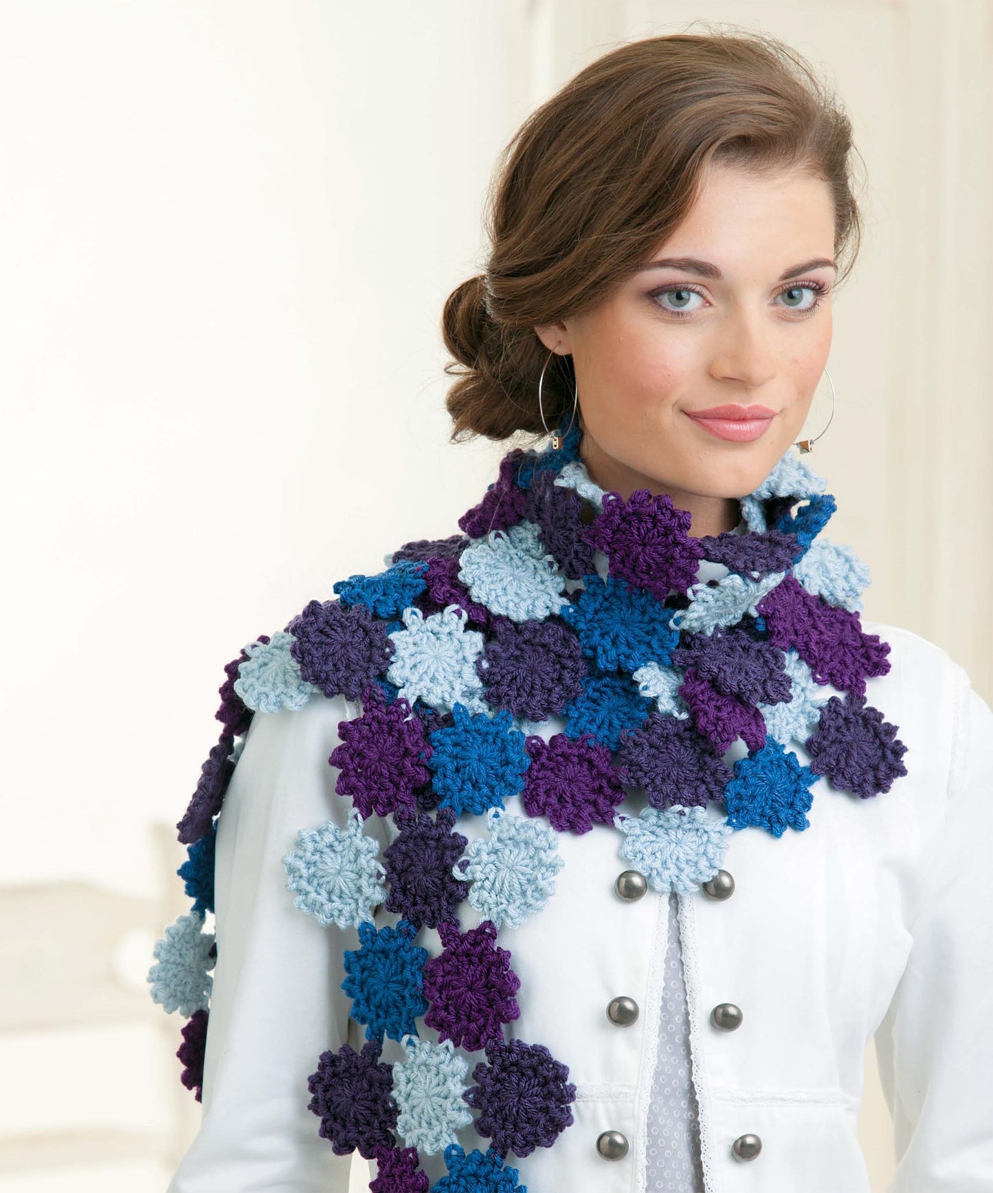 A woman with brown hair in an updo, smiling softly indoors, wears a white coat and a crocheted scarf of blue, purple, and light blue flower motifs—perfect inspiration for Leisure Arts' Beginner's Guide to Crochet Motifs.