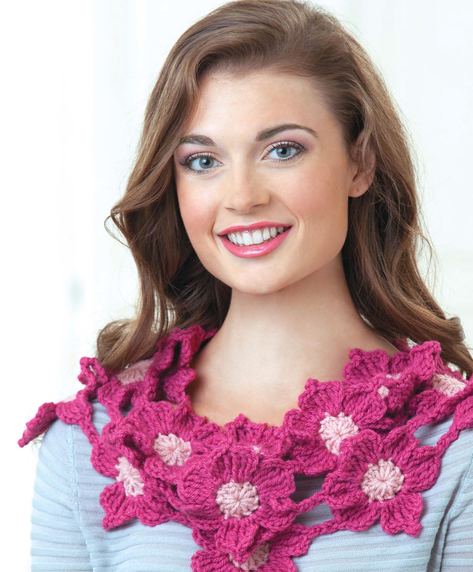 A woman with long brown hair smiles, wearing a light purple top and a chunky pink and mauve crocheted flower scarf—perfect inspiration for projects from Leisure Arts’ Beginner's Guide to Crochet Motifs.