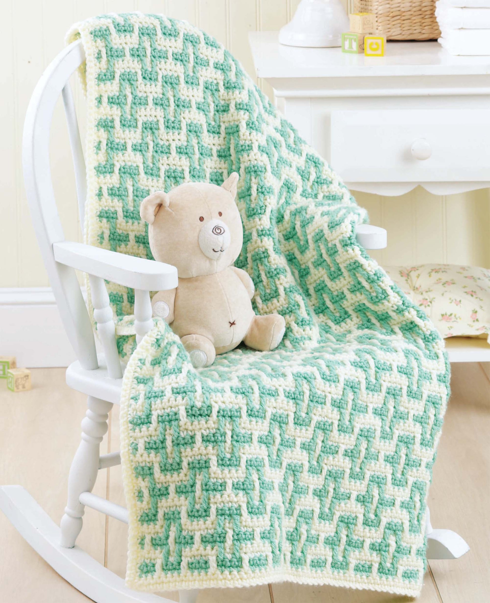 A soft beige teddy bear sits on a white rocking chair draped with a mosaic crochet blanket made using techniques from the Beginner's Guide to Crochet Color Work by Leisure Arts. A white dresser and pastel decor appear in the background.