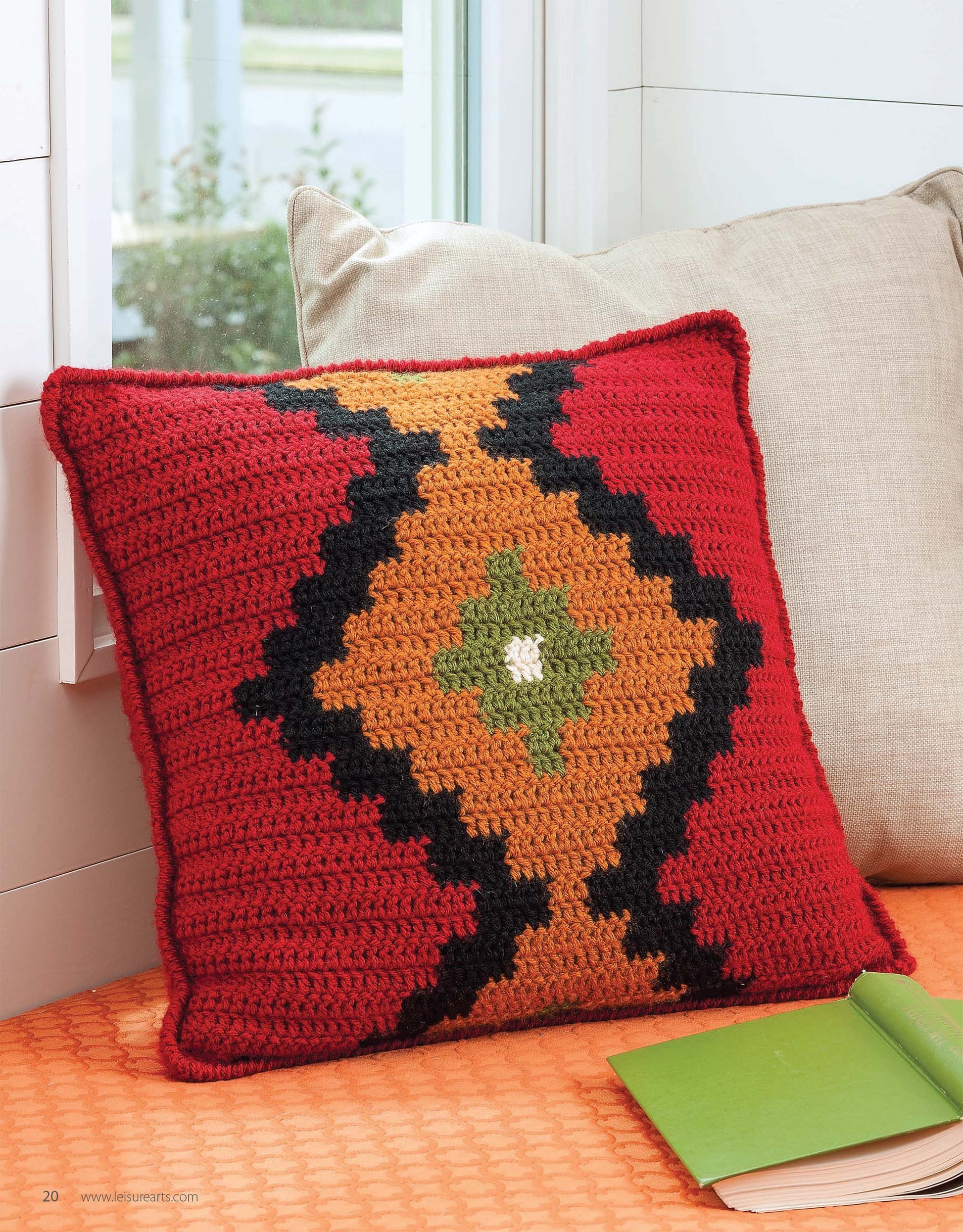 A mosaic crochet pillow, inspired by the Beginner's Guide to Crochet Color Work from Leisure Arts, features a red background and diamond-shaped geometric patterns in black, orange, green, and white on an orange textured surface beside beige cushions.