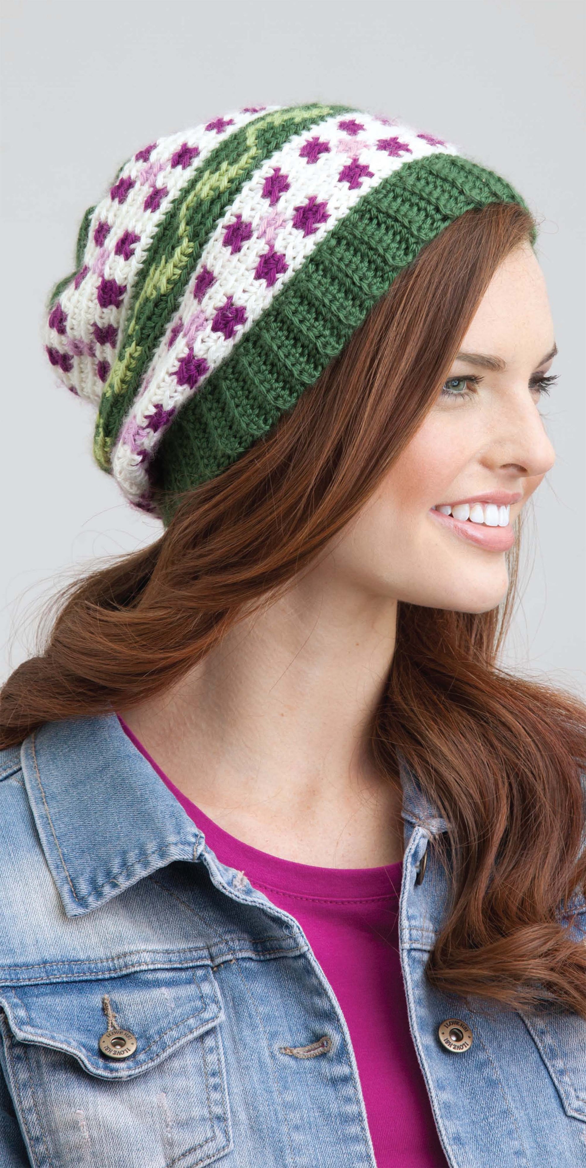 A woman smiles while wearing a green, white, and purple fair isle-inspired beanie she made using the "Beginner's Guide to Crochet Color Work" from Leisure Arts. She has long reddish-brown hair and wears a denim jacket over a magenta shirt against a light gray background.