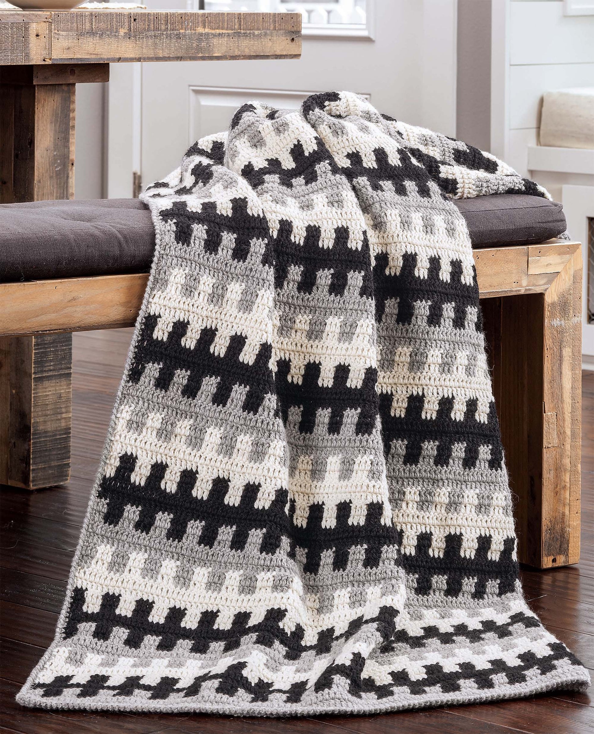 The "Beginner's Guide to Crochet Color Work" by Leisure Arts features a mosaic crochet geometric blanket in black, gray, and cream, beautifully displayed on a rustic wooden bench in a sunlit, cozy space.