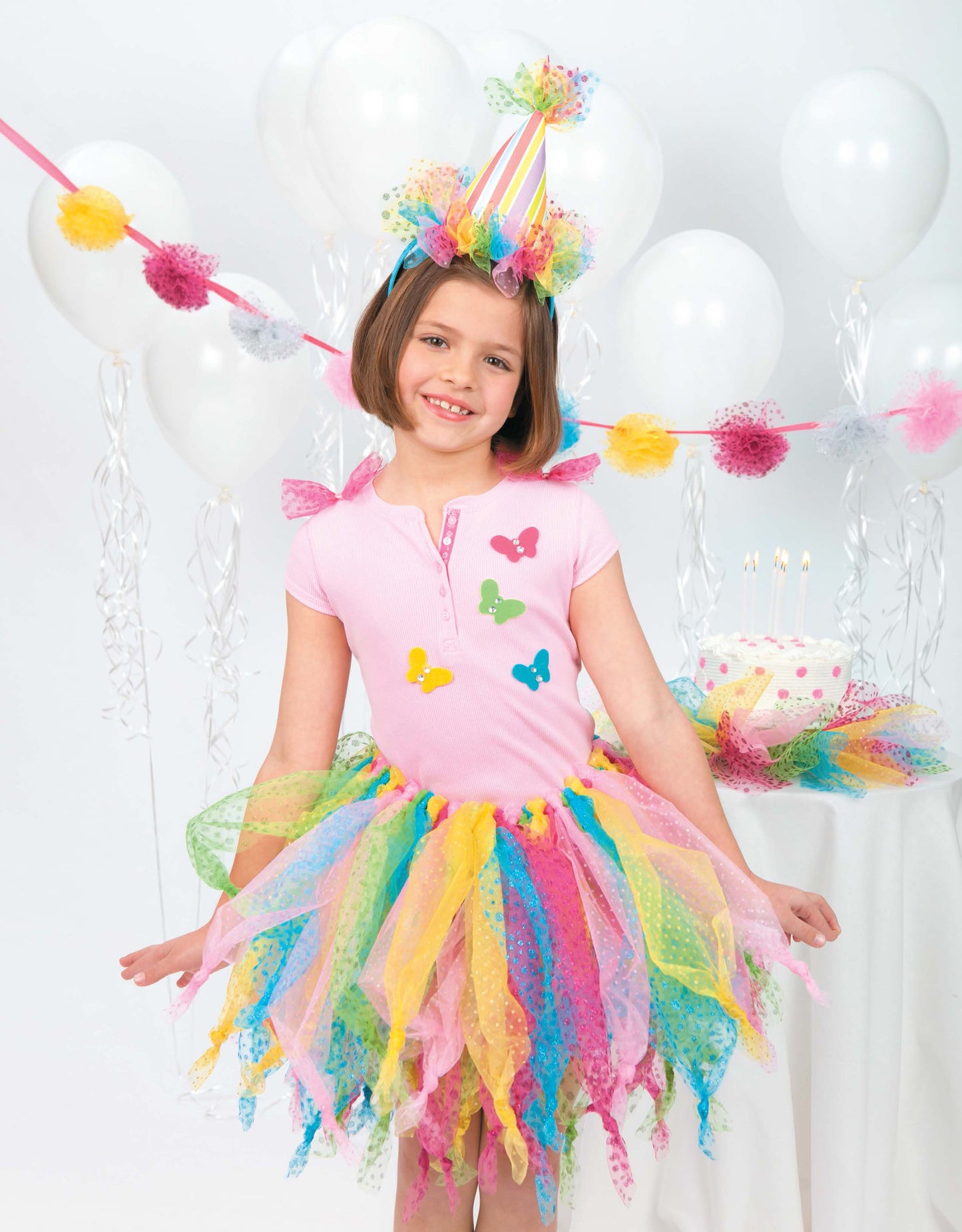 A smiling girl in a "A Tutu for Every Occasion" dress by Leisure Arts stands before white balloons and a birthday cake, ready to celebrate.