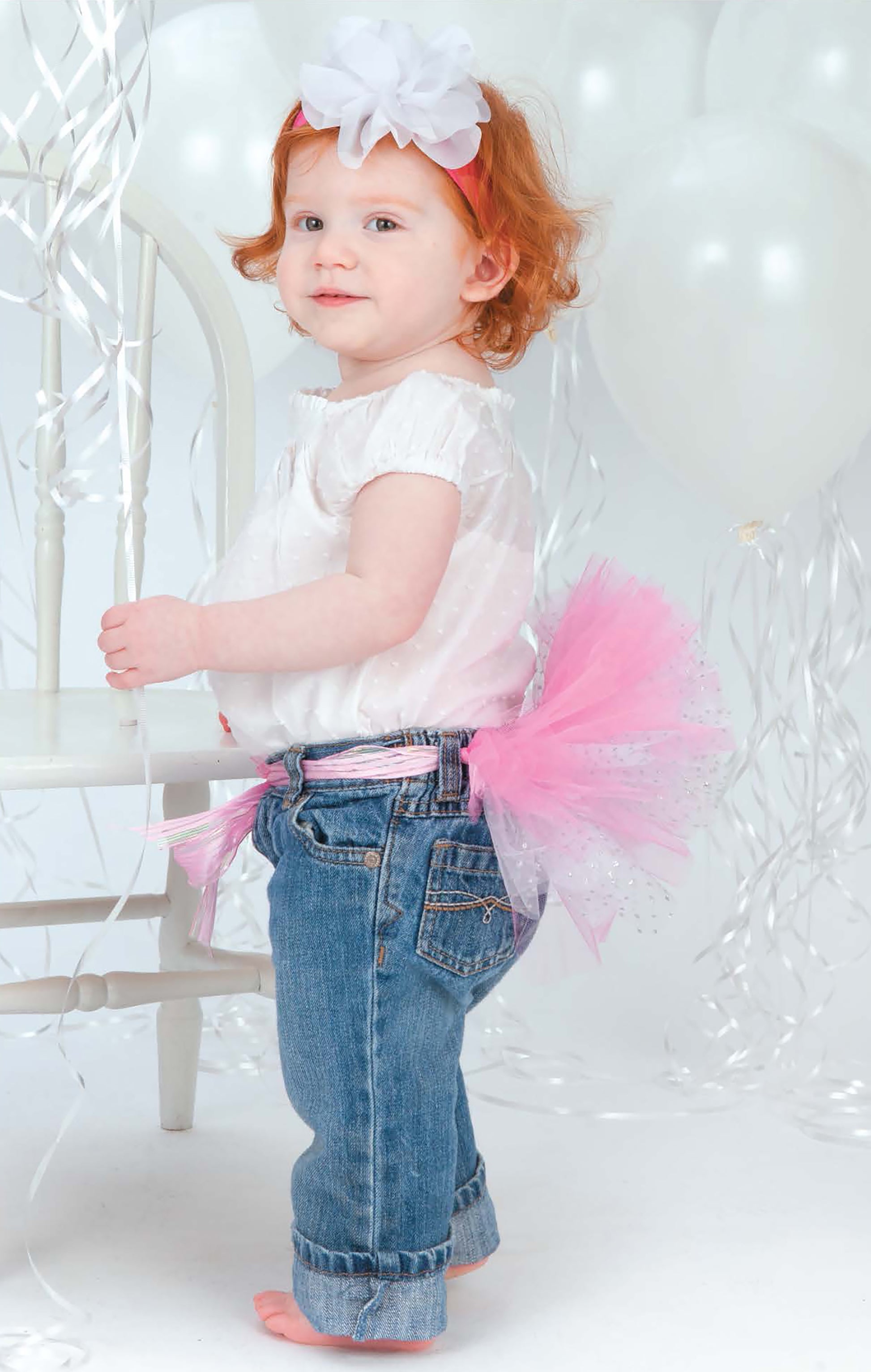 A toddler in a white top, jeans, and the Leisure Arts "A Tutu for Every Occasion" pink tutu stands barefoot by a white chair amid balloons and streamers, wearing a white flower headband.