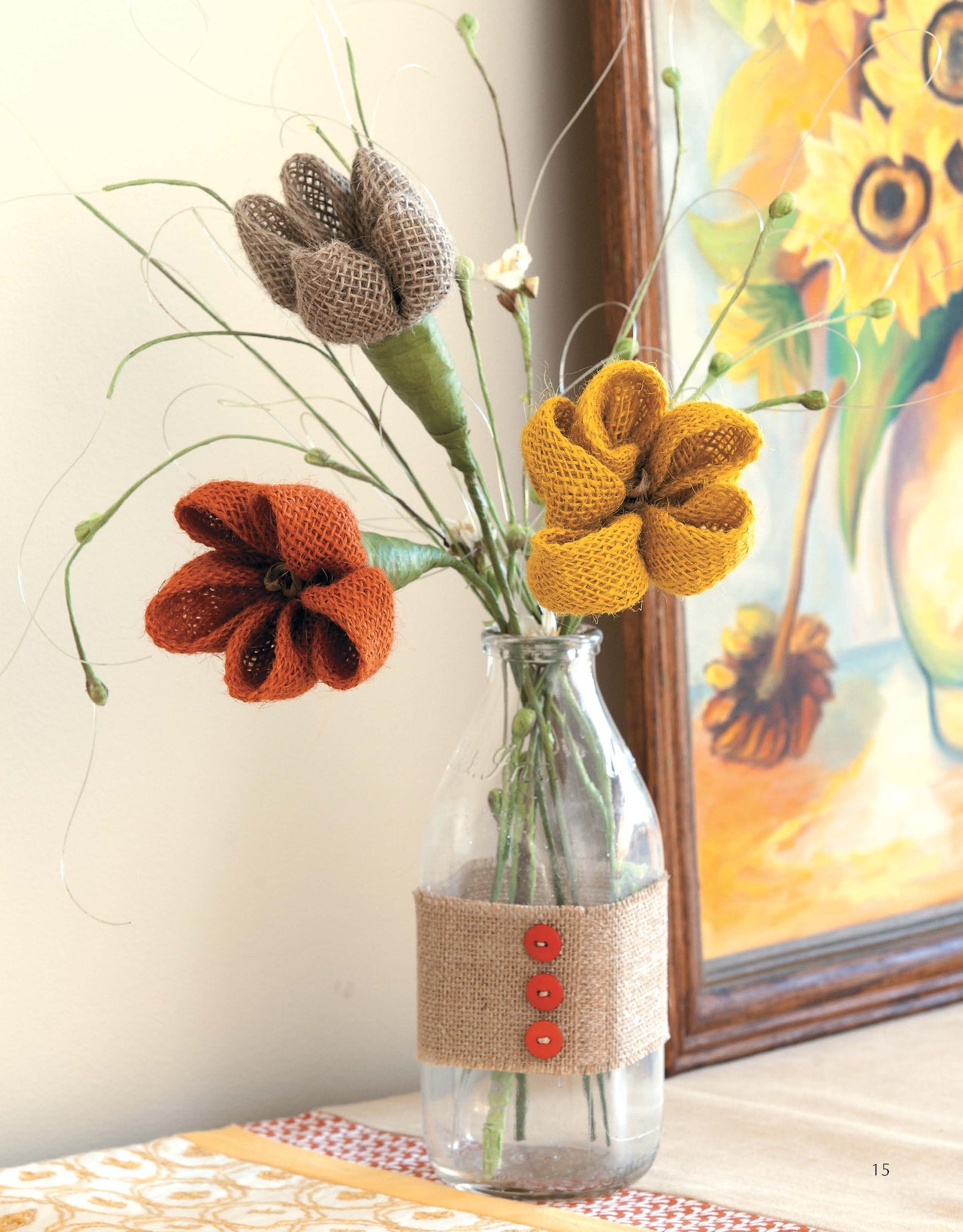 Leisure Arts' "Decorate with Ribbon" features a glass bottle wrapped in burlap and orange buttons, holding three burlap flowers in orange, yellow, and brown—ideal for beginner home decor and paired with a sunflower painting.