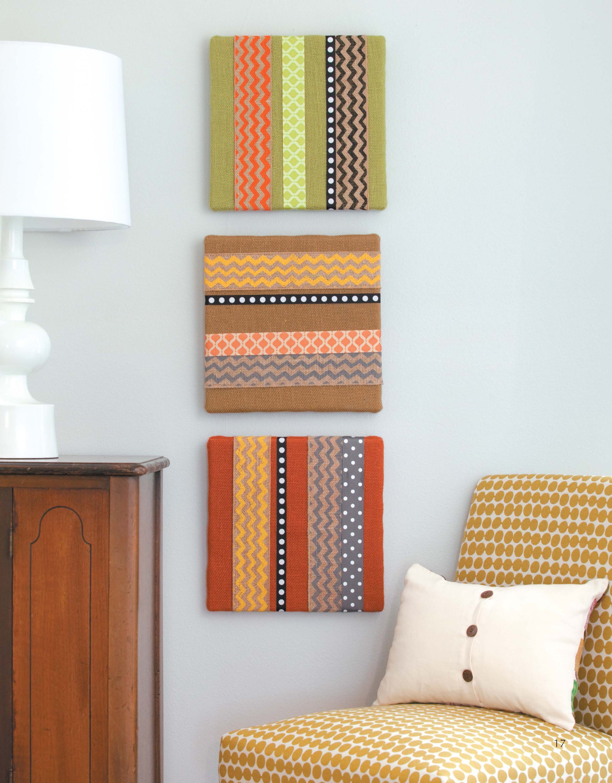 Decorate with Ribbon by Leisure Arts features three square fabric wall arts with vibrant zigzag and dot patterns, hung vertically above a cabinet next to a yellow chair, white pillow, and lamp—ideal for beginner home decor or no-sew projects.