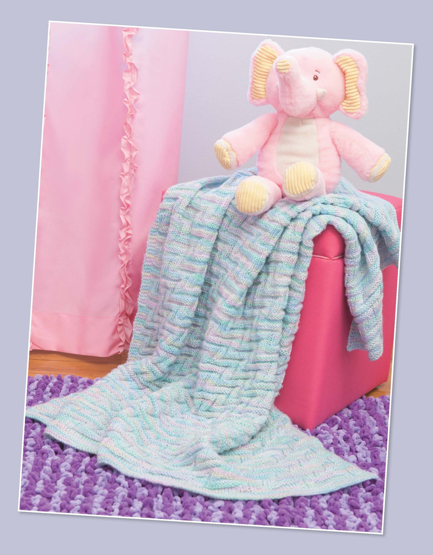A pink plush elephant sits on a pink ottoman draped with a pastel blanket from Leisure Arts' "Projects for Baby Made with the Knook." A purple textured rug covers the floor, and a pink ruffled curtain hangs in the background.