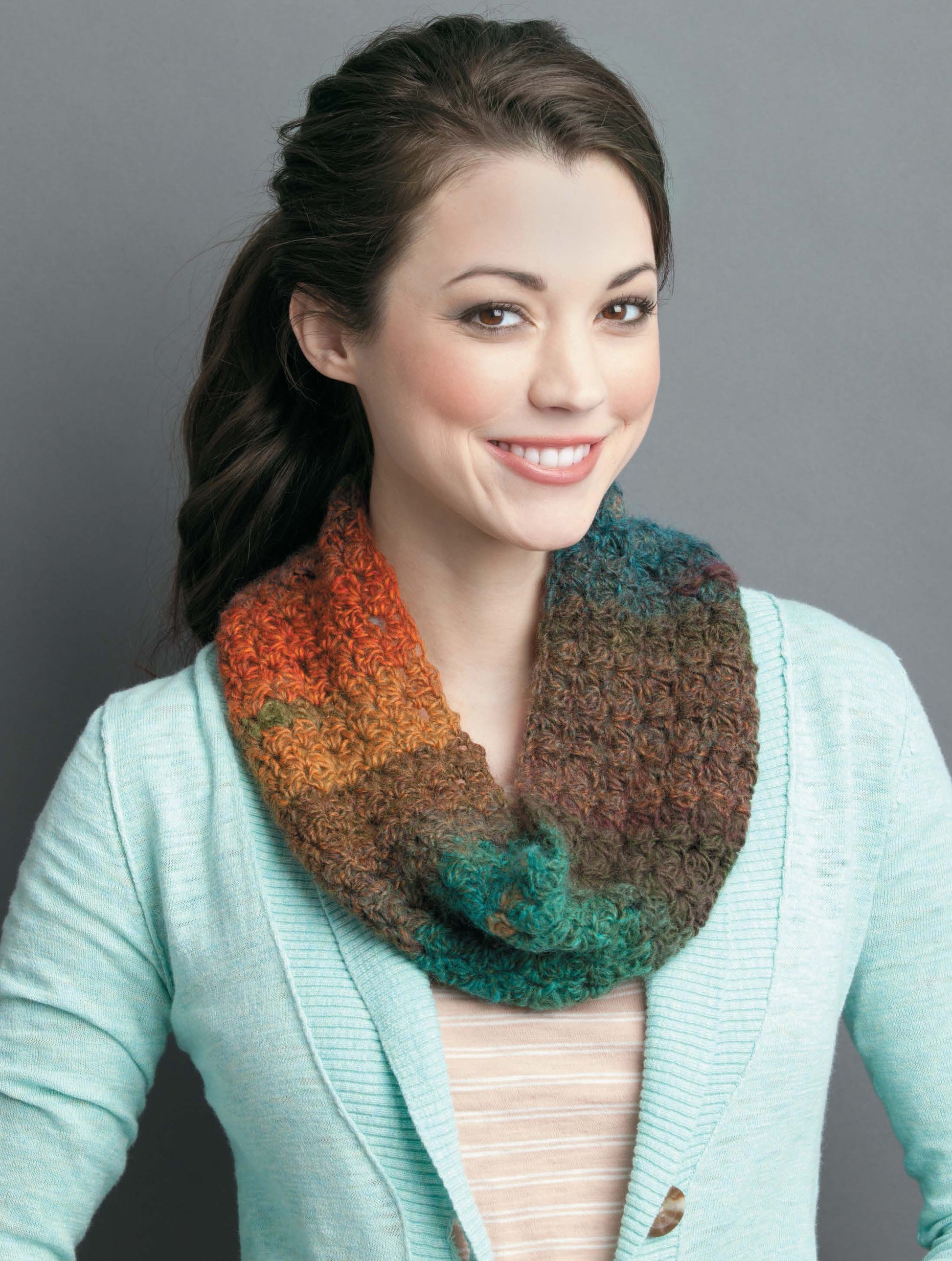 A smiling woman with long brown hair in a ponytail wears a light blue cardigan and multicolored infinity scarf, showcasing inspiration from Leisure Arts' "Crochet Scarves & Cowls" Digital Download, against a plain gray background.