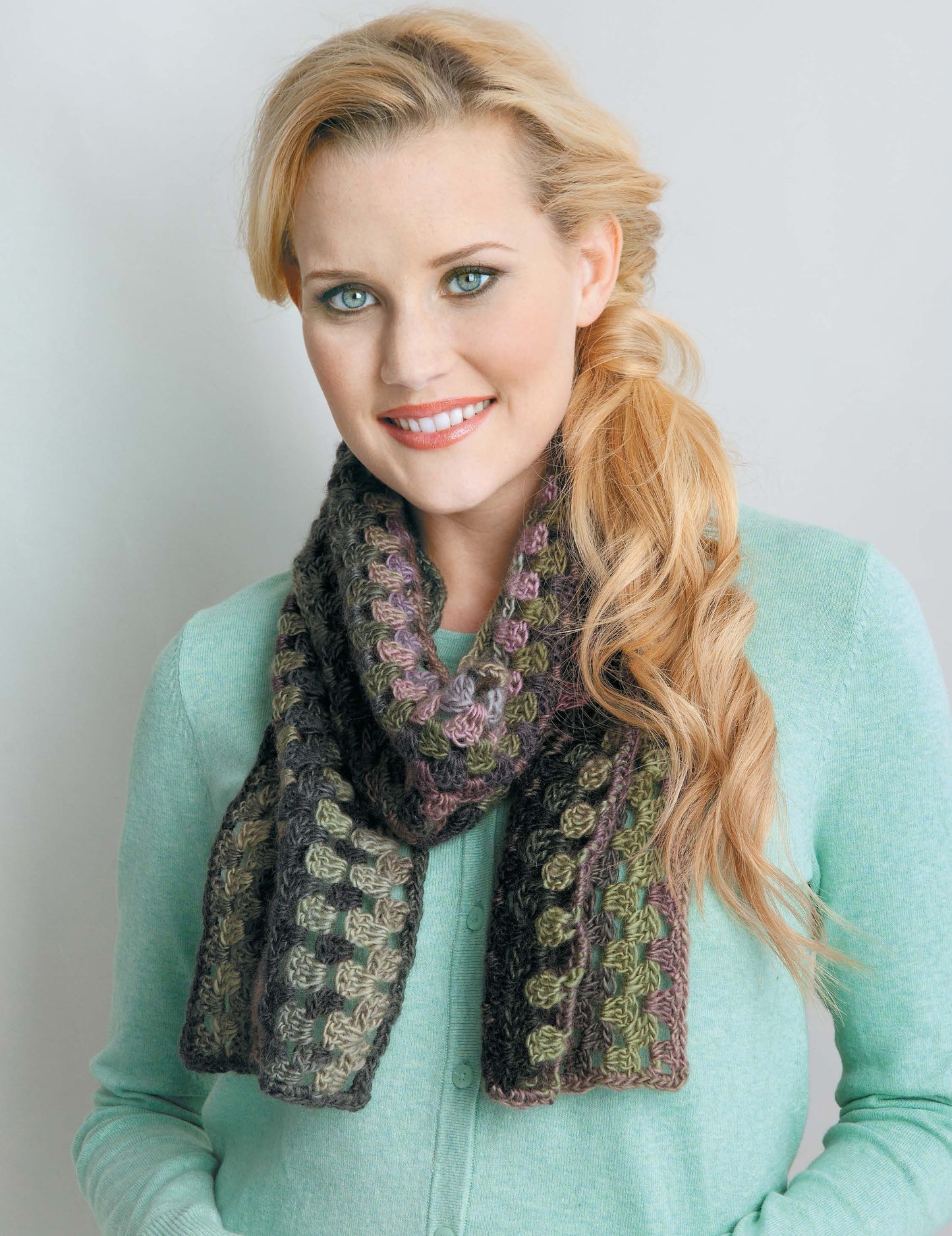 A smiling woman in a mint green top models a chunky green and brown crochet scarf—an ideal accessory from the Crochet Scarves & Cowls Digital Download by Leisure Arts—against a light gray background.