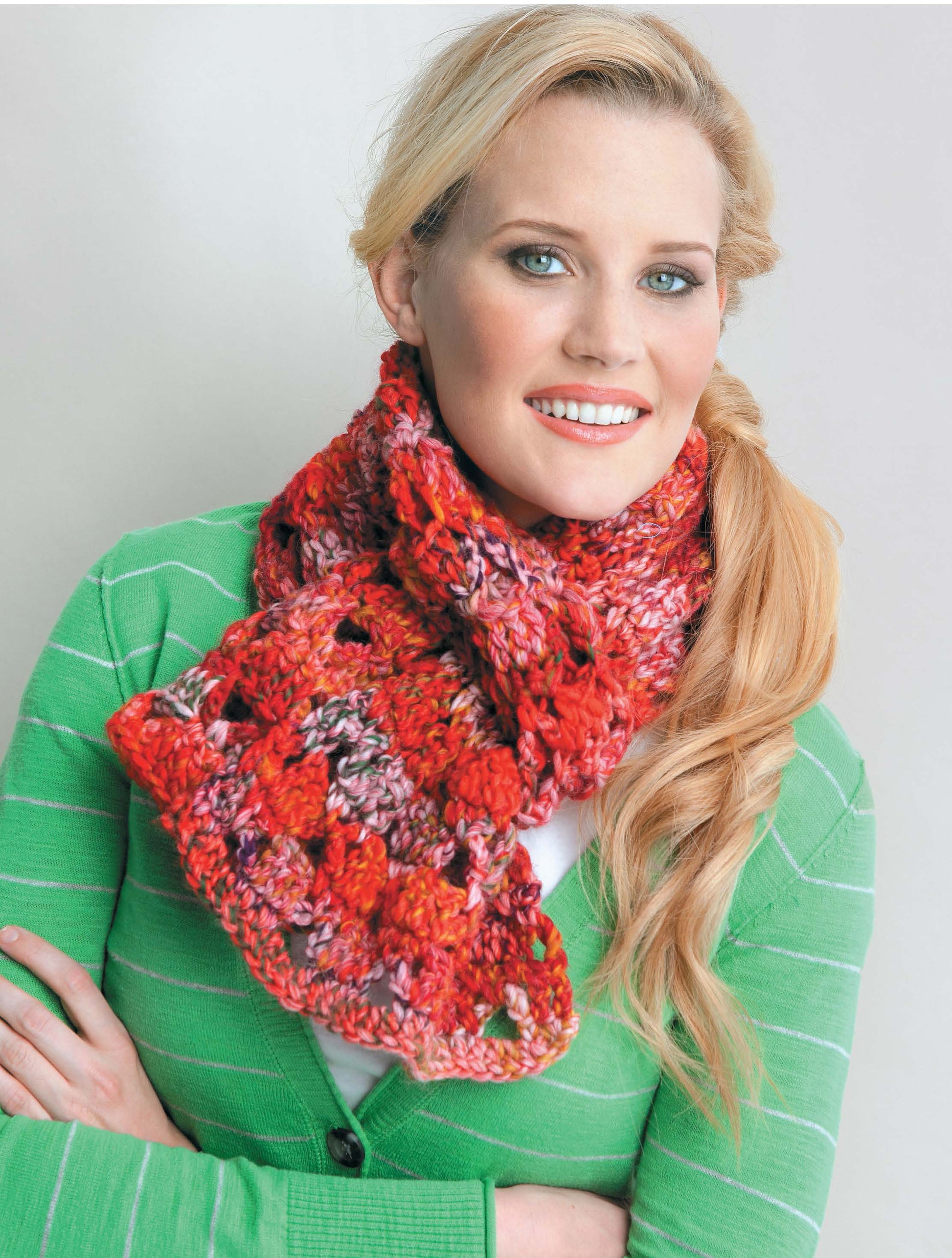A smiling woman in a green striped sweater and a chunky red and pink crocheted scarf showcases fresh ideas from the Crochet Scarves & Cowls Digital Download by Leisure Arts—perfect for beginners or anyone seeking new scarf inspiration.